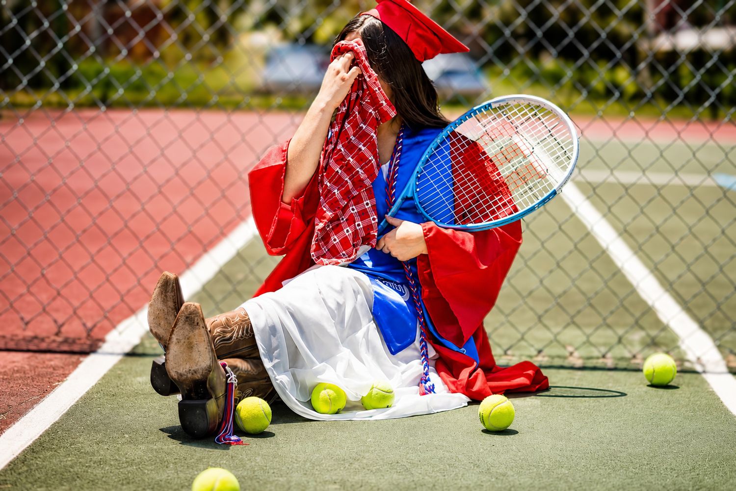 Woman in red and white dress sits on tennis court holding racket surrounded by yellow tennis balls.