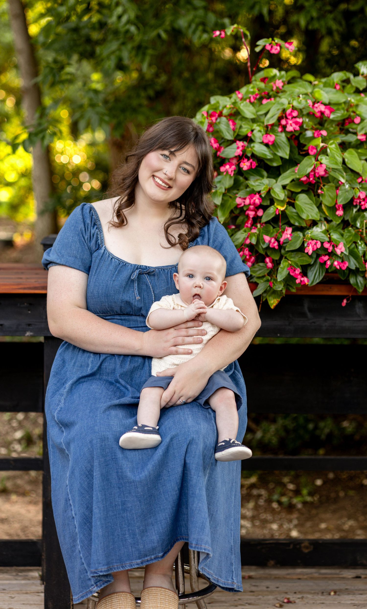 A smiling young woman in a blue denim dress holds a cute baby while sitting in front of pink flowers outdoors.