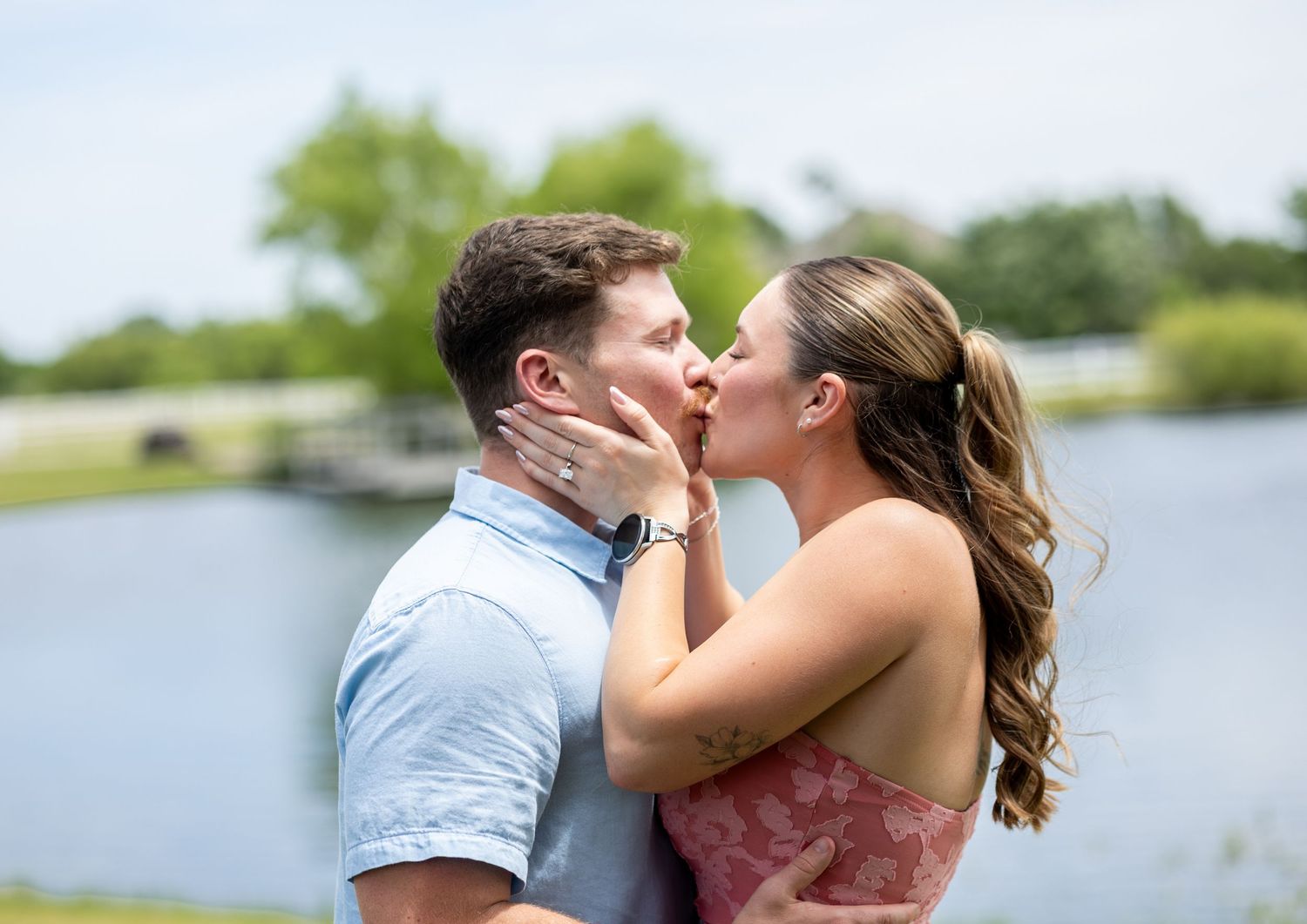 Romantic couple sharing a kiss outdoors near a serene lake on a sunny day.