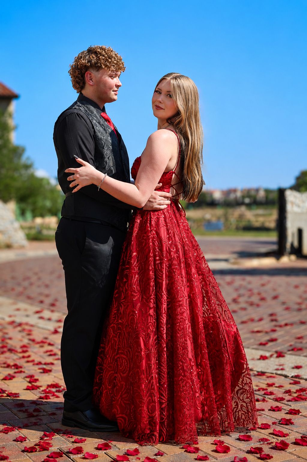 A young couple in prom attire, man in black suit and woman in red lace gown, pose on a rose petal-covered path.