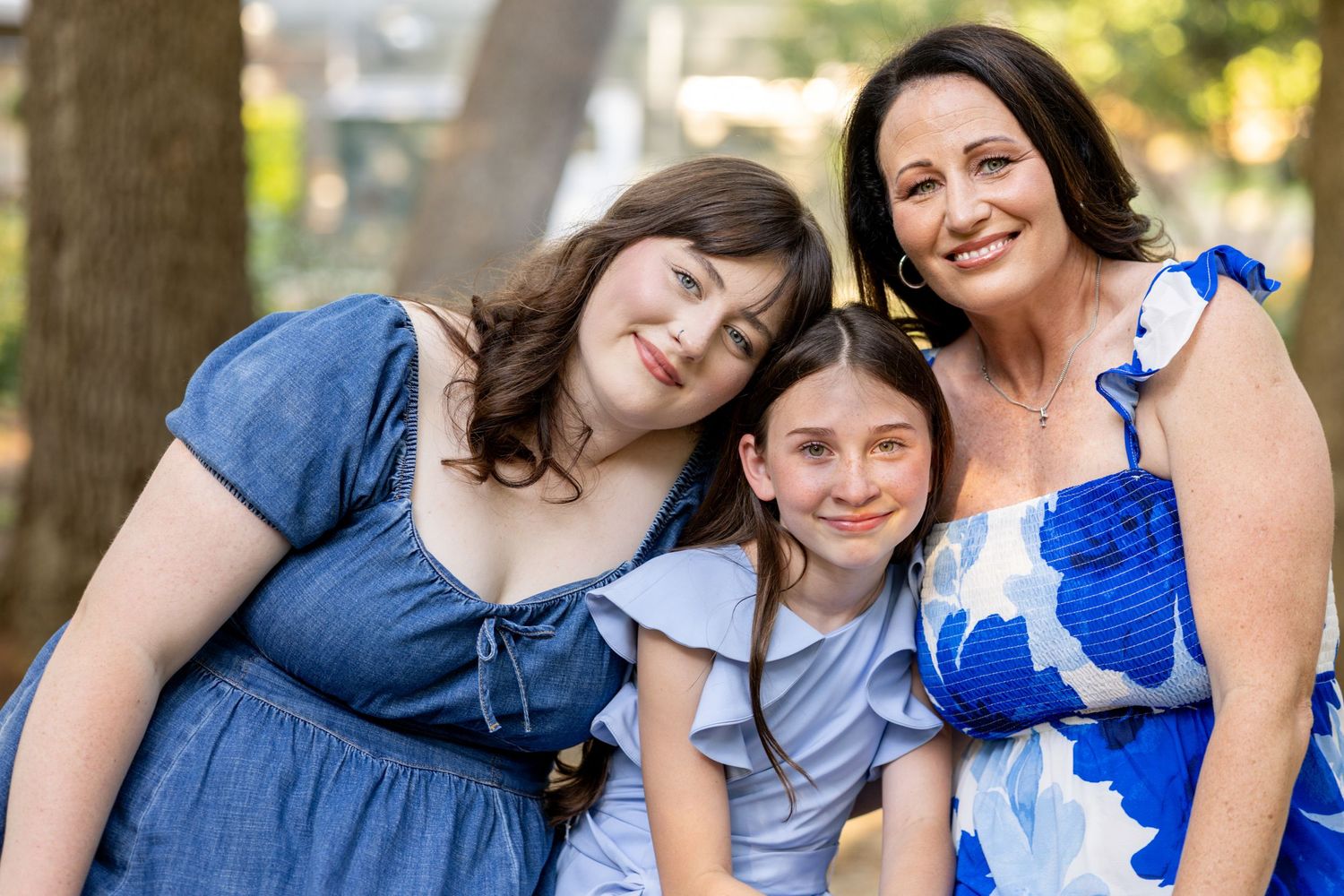 A mother smiles with her two daughters outdoors, all wearing blue outfits, in a warm family portrait.