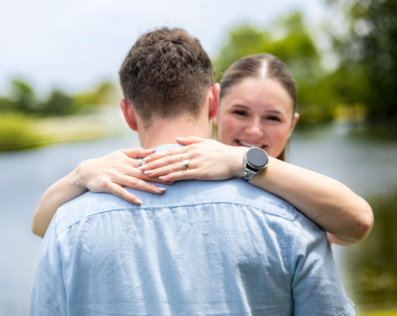 A smiling woman with a watch hugs a man in a blue shirt outdoors near a serene pond.