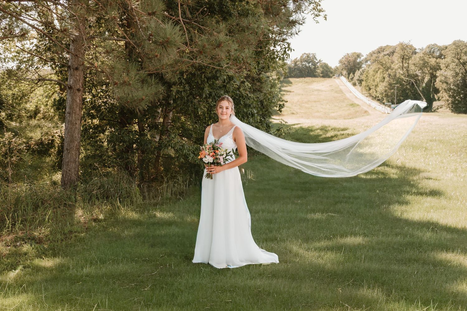 Bride outside a grassy field near a ski slope with veil blowing in the wind in Minnesota