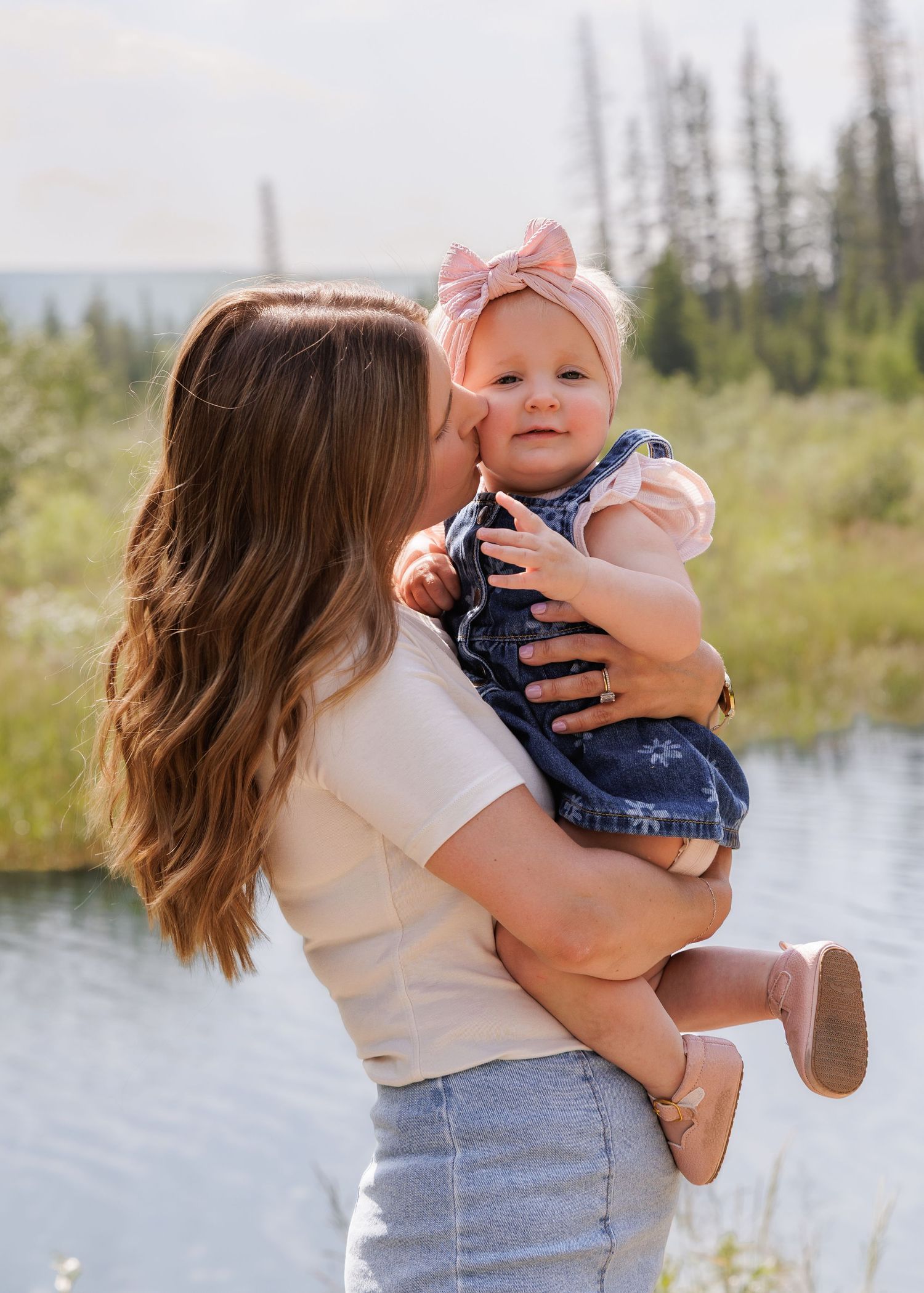 A mother kisses her baby girl wearing a pink bow and denim overalls near a scenic lake surrounded by trees.