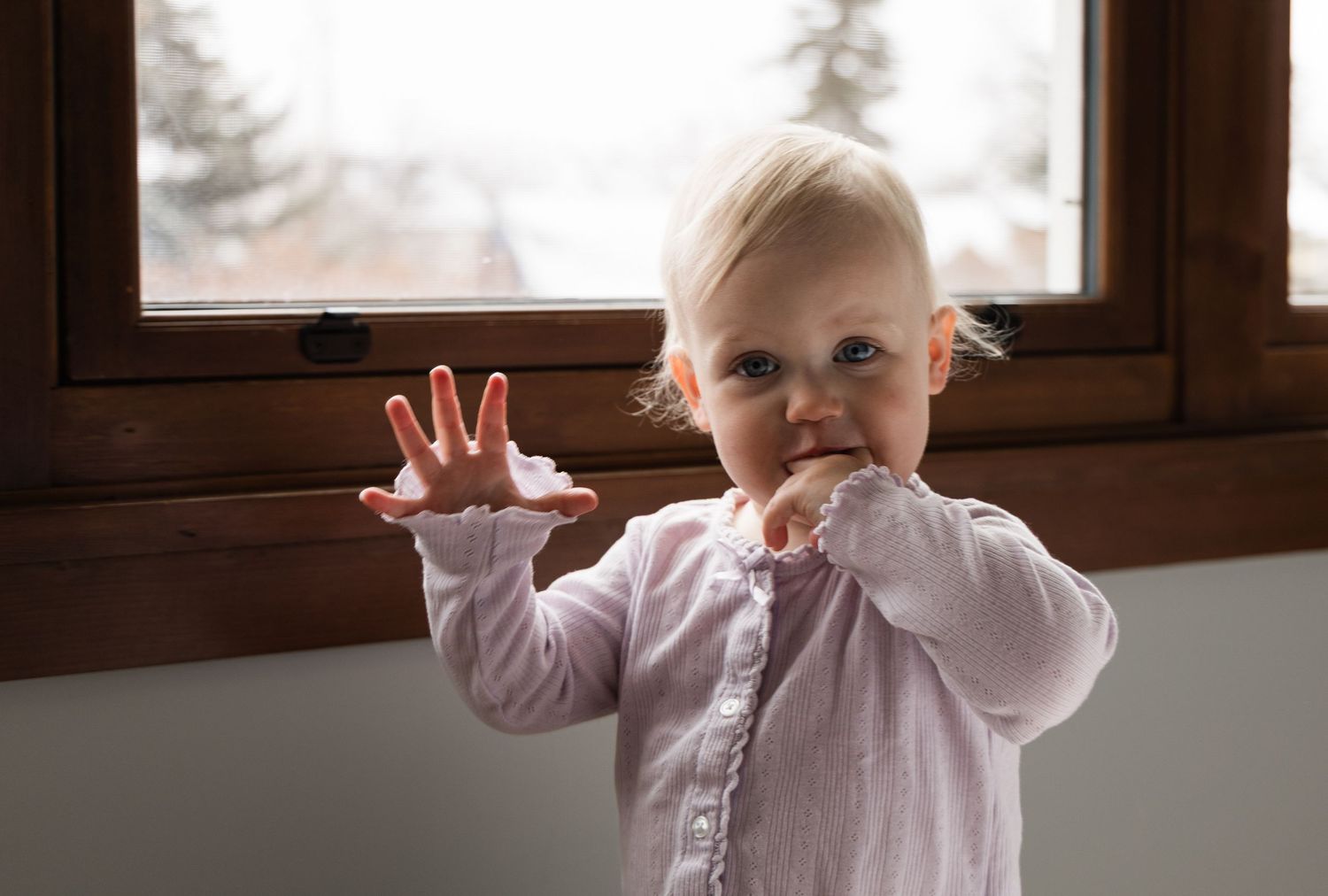 Adorable blonde toddler in pink cardigan standing by window, reaching out with hand toward camera.