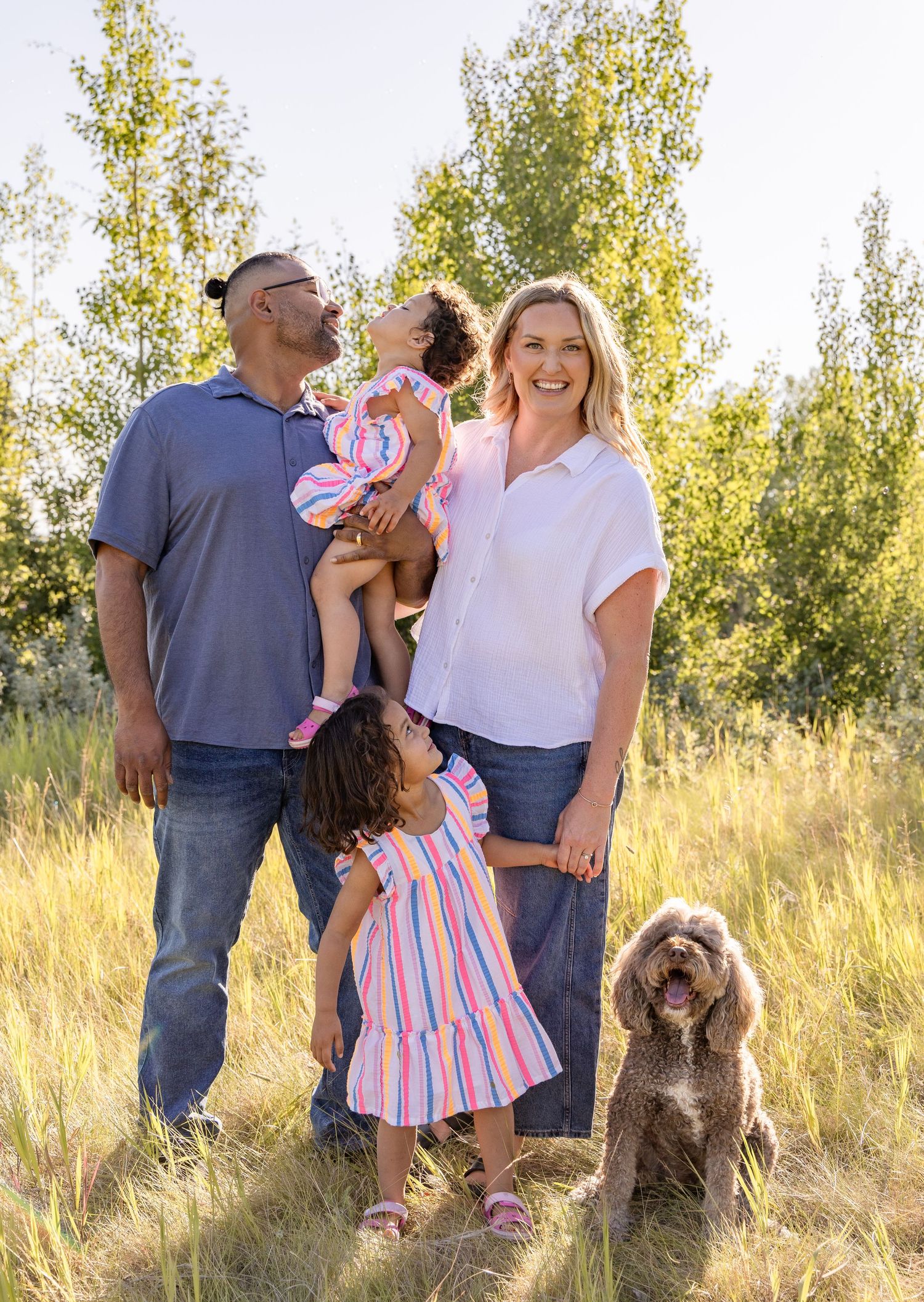 Family of four with a fluffy brown dog posing outdoors in a sunny meadow with green trees in the background.