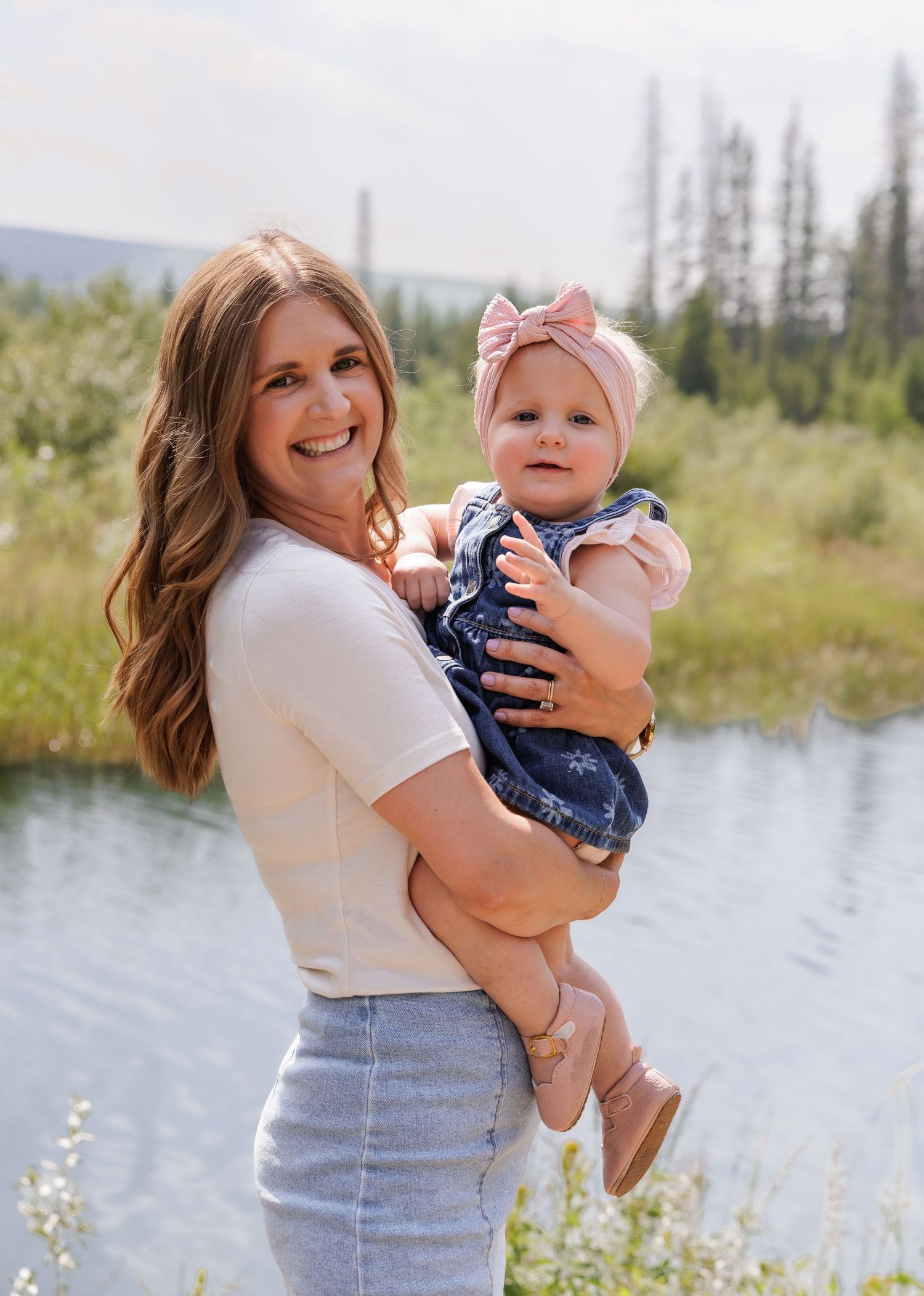 A smiling woman holds a baby girl in a pink bow near a serene mountain lake surrounded by evergreen trees.