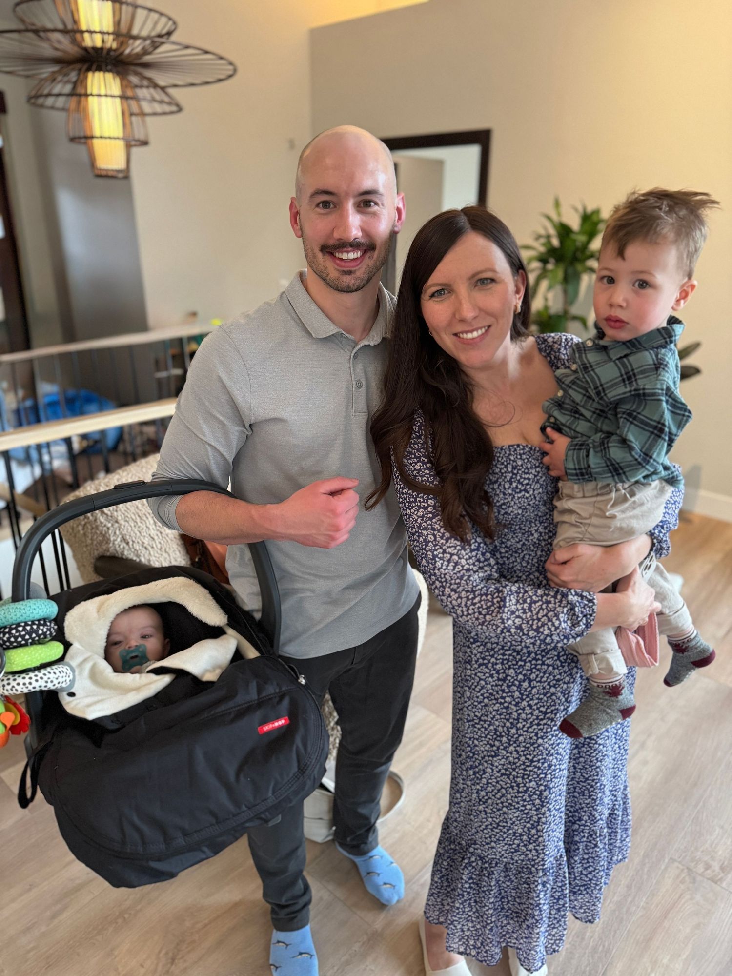 Happy family of four posing indoors with a newborn in a car seat and a toddler.