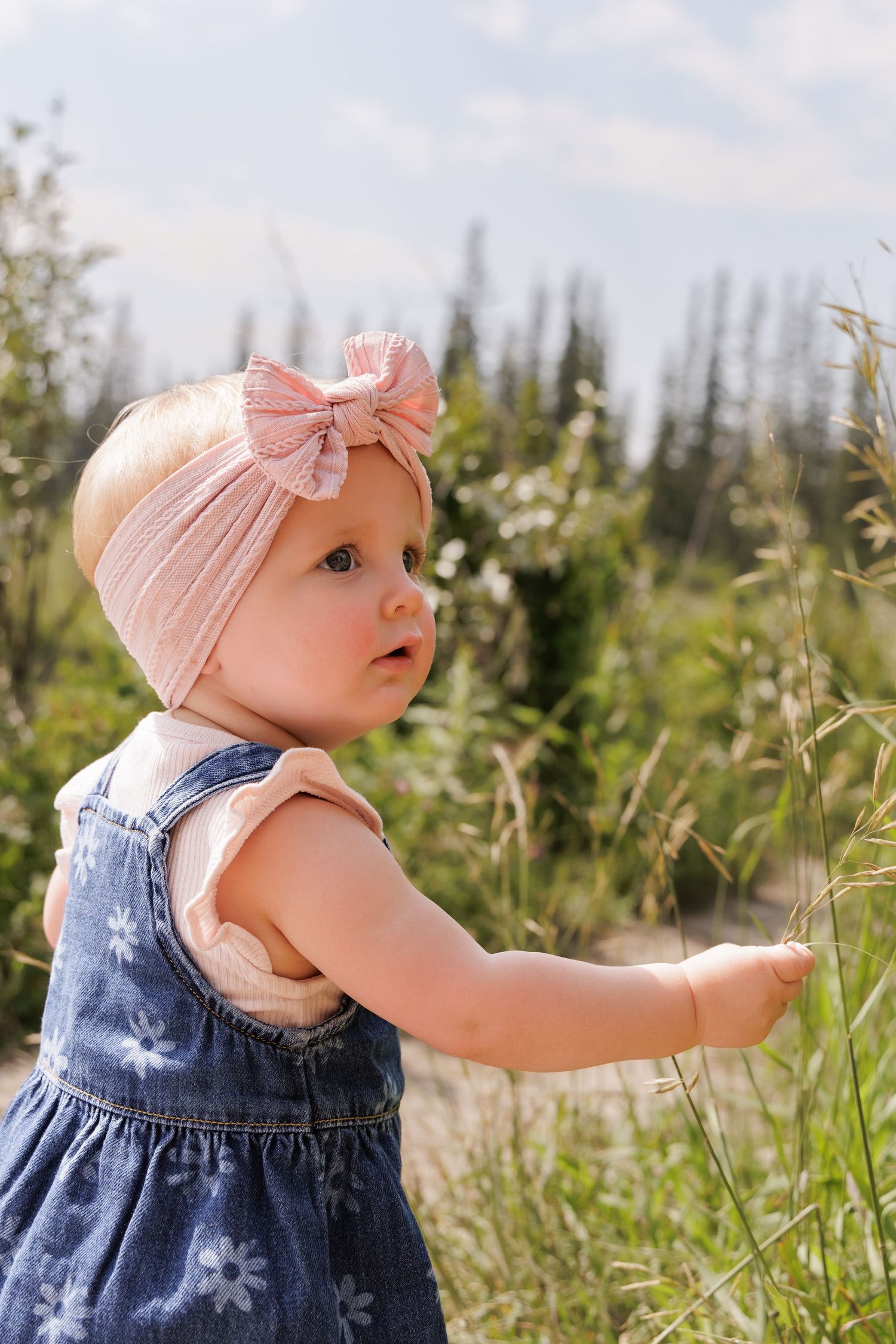 Baby girl in denim floral dress and pink bow headband touching tall grass in a sunny outdoor field.