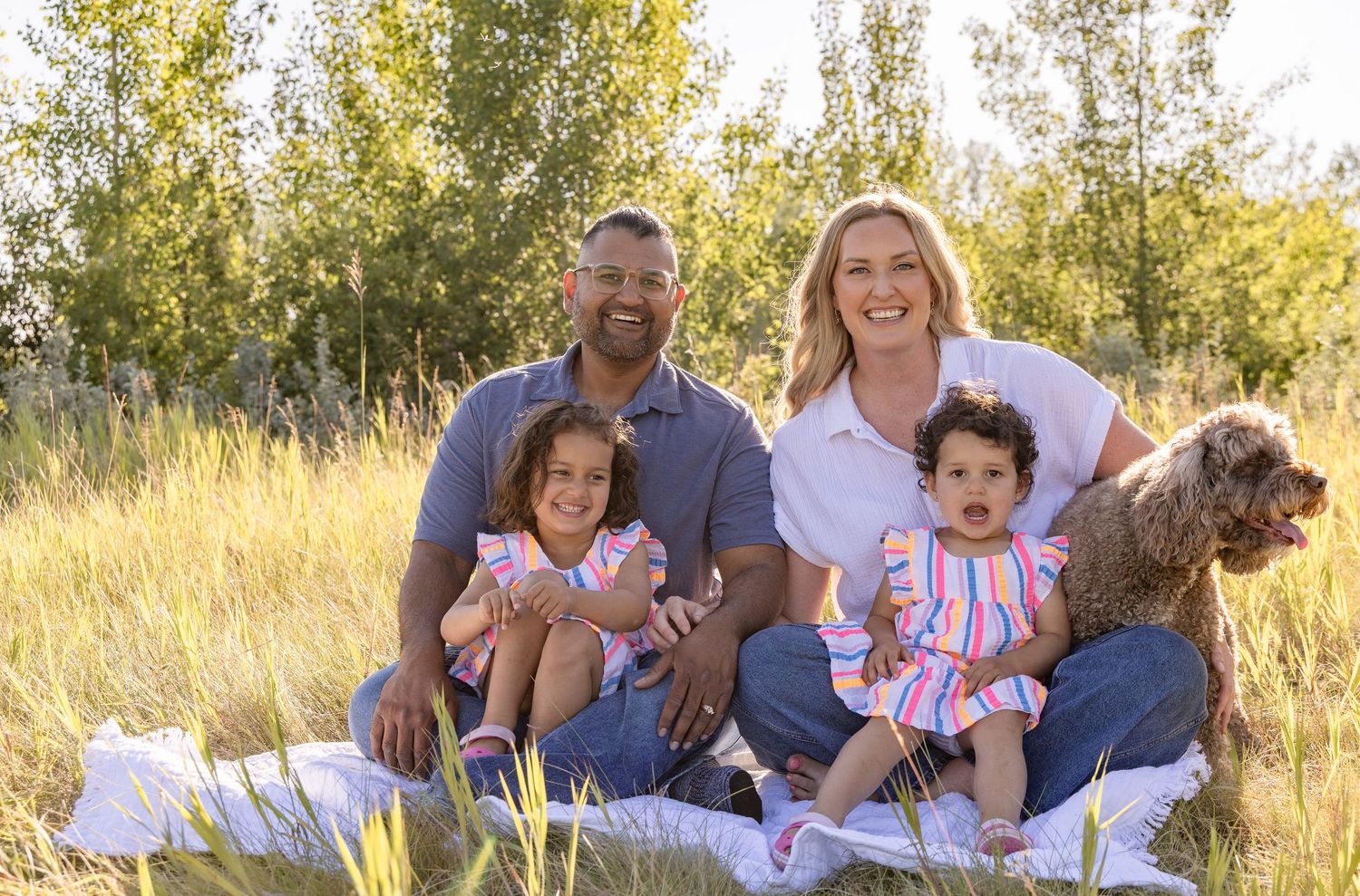 Family of four with dog sitting in sunny meadow, parents smiling with two young daughters in colorful outfits.