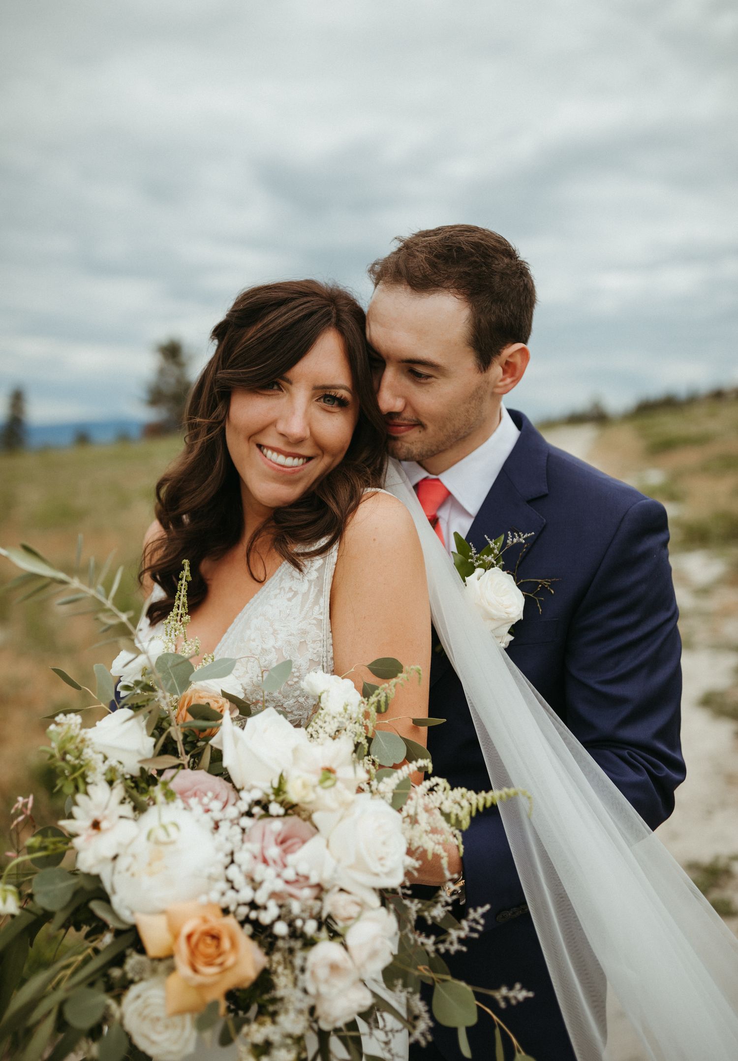 A bride holding a lush floral bouquet leans into her groom in a navy suit outdoors on their wedding day.