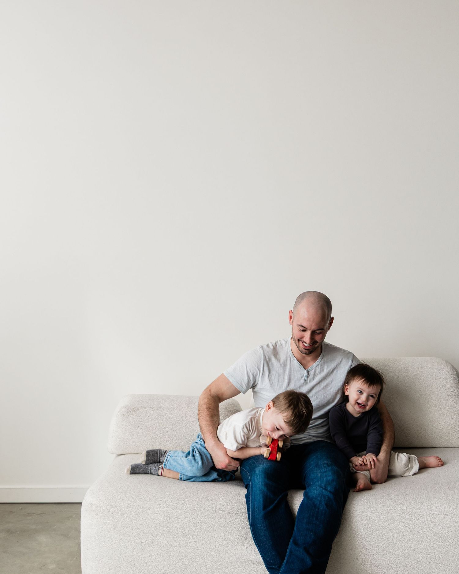 A bald father sits on a beige sofa with two young children, smiling warmly in a bright, minimalist living room.