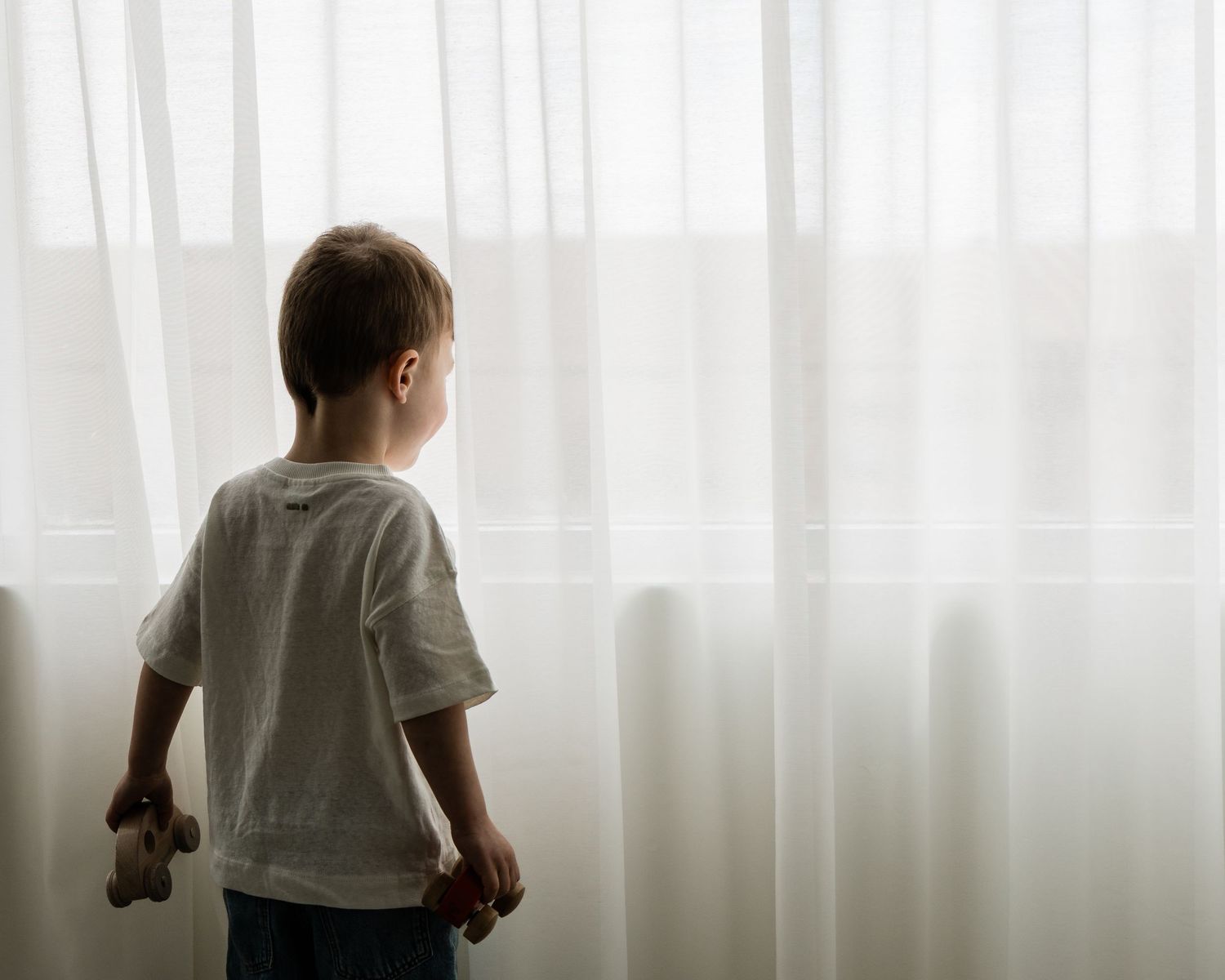 Young boy in white t-shirt standing alone at a bright window holding a small stuffed animal toy.
