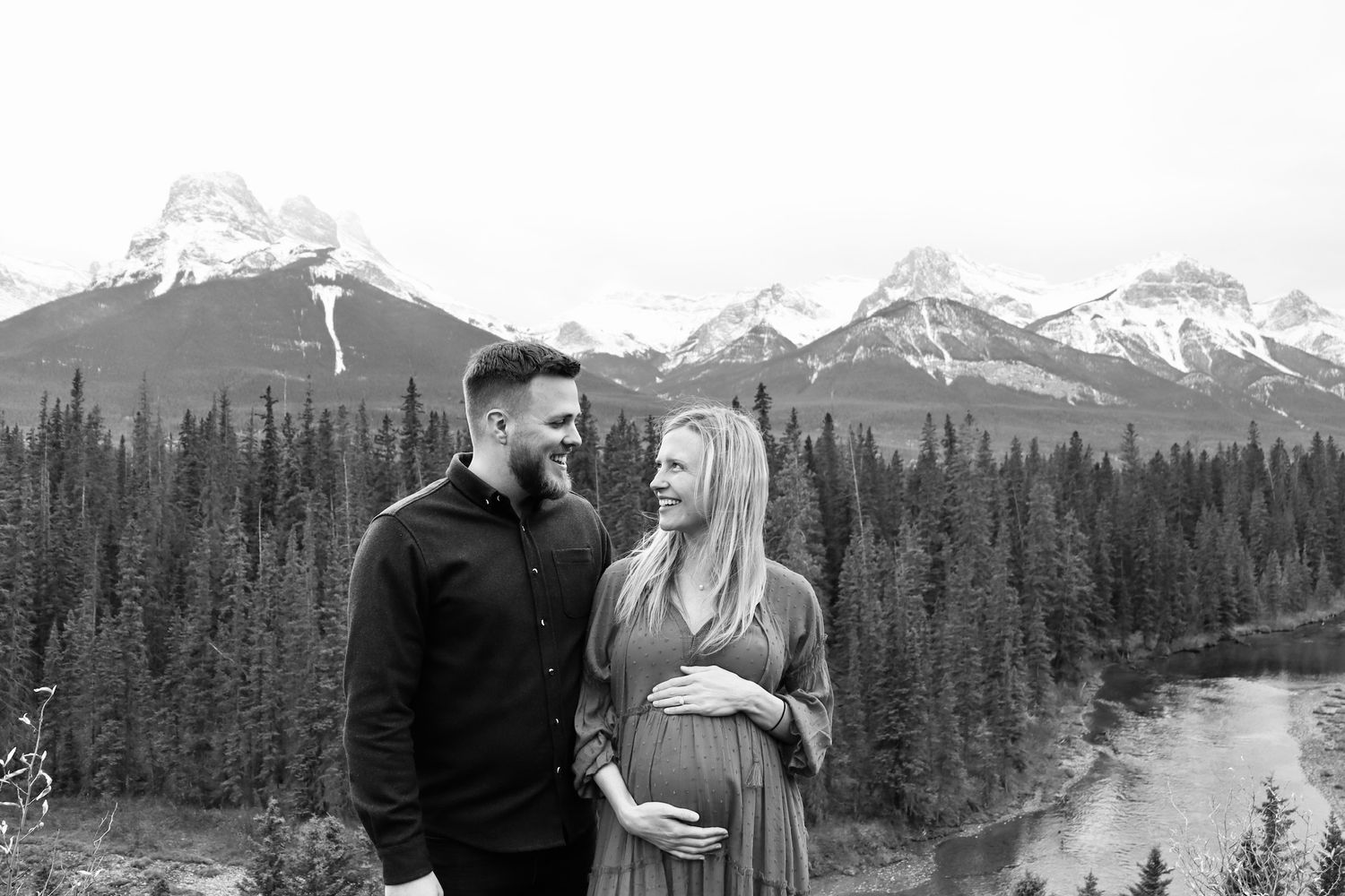 Couple posing for maternity photo with stunning mountain and river backdrop in black and white.