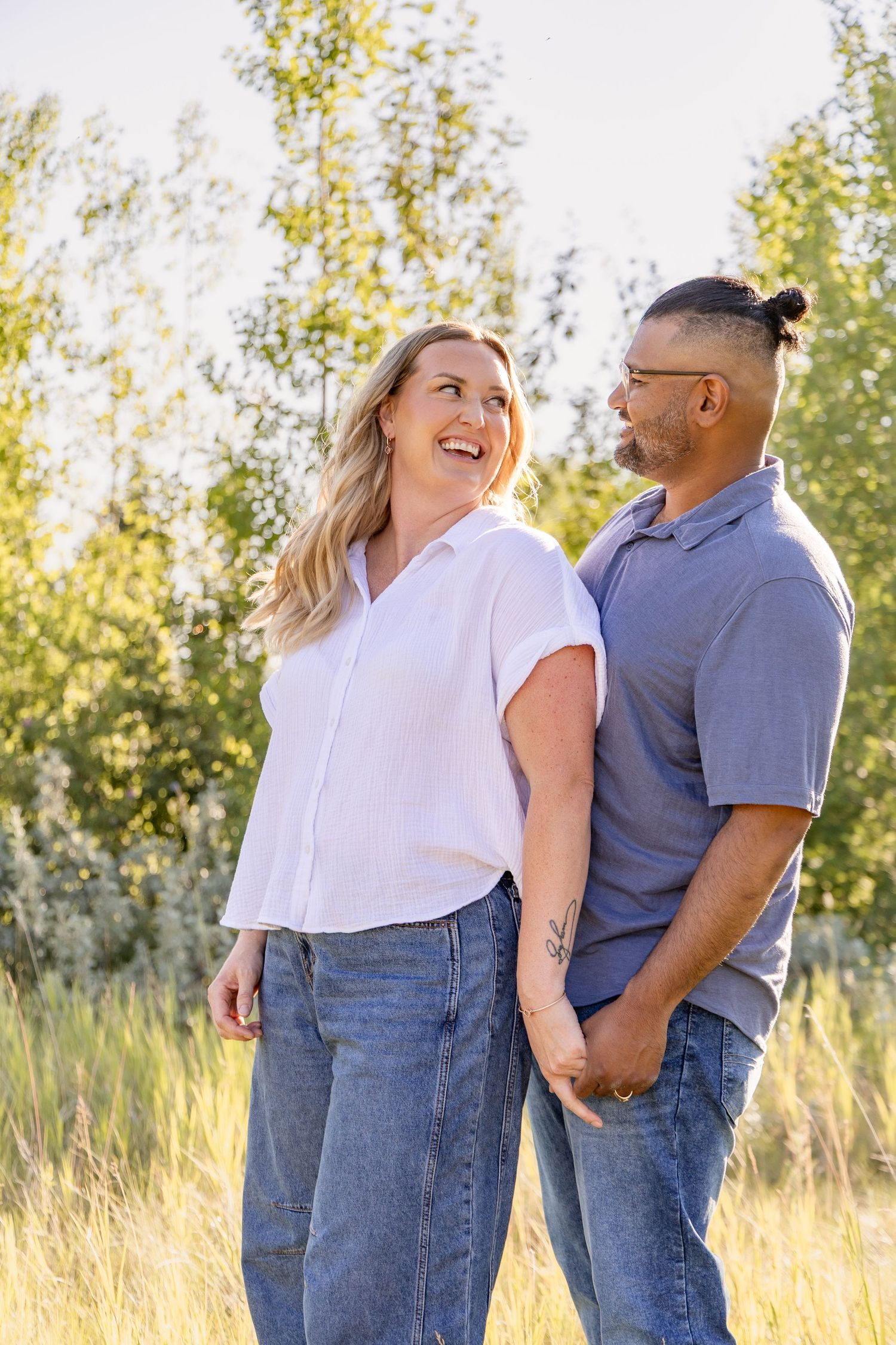 A smiling couple holds hands in a sunny field, the woman in a white shirt and the man in a blue polo, both wearing jeans.