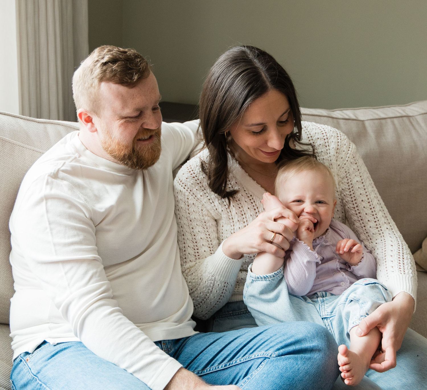 Loving parents sitting on couch smiling at their happy laughing baby girl.