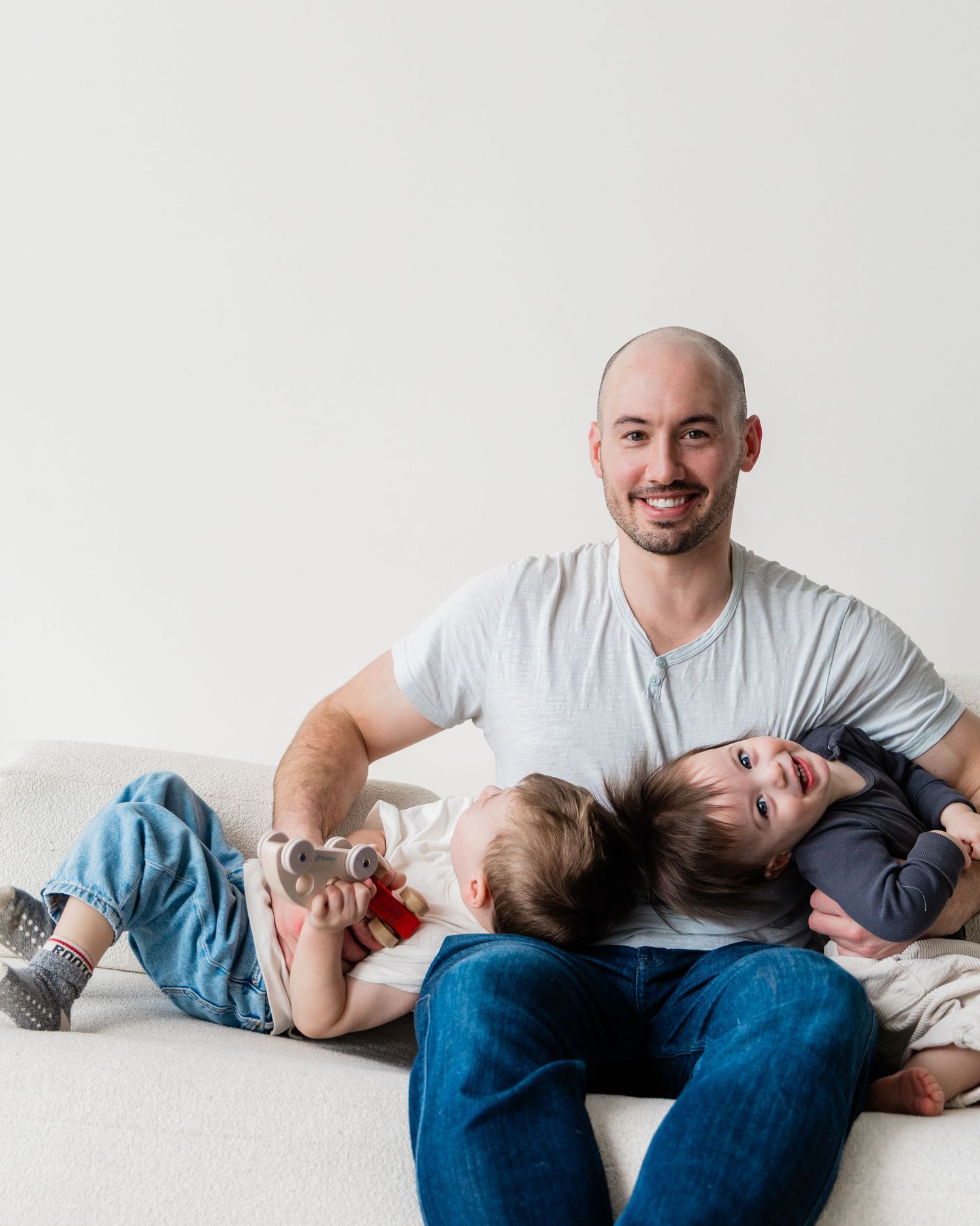 Smiling bald father relaxing on a white couch with his two young children lying across his lap.