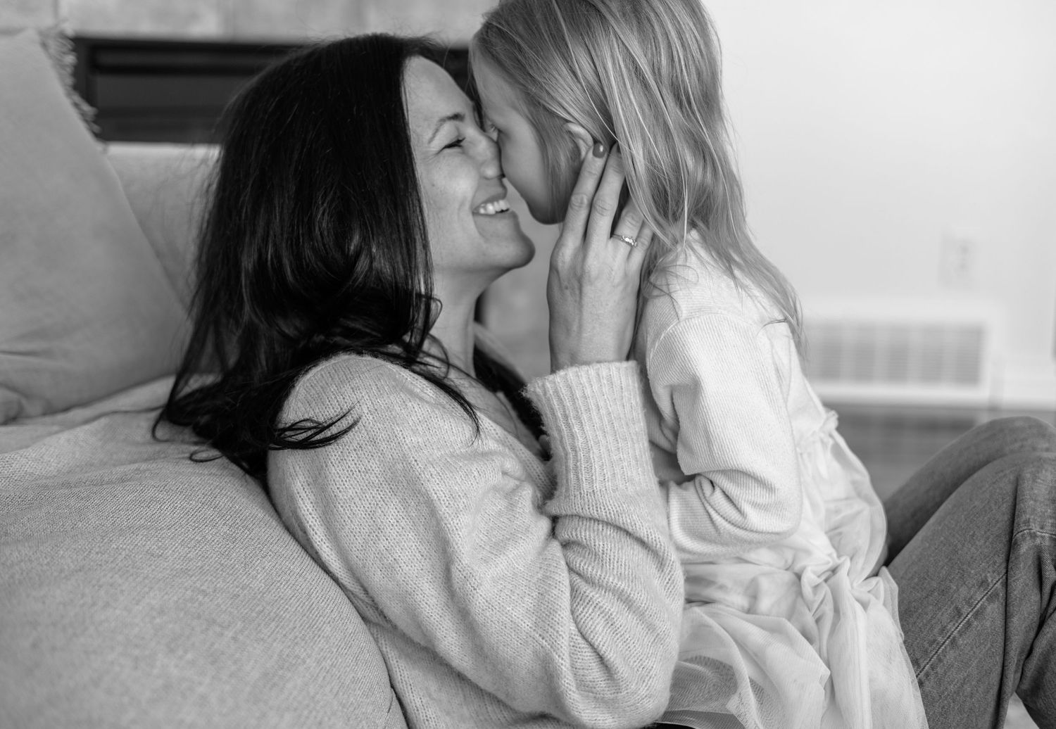 Black and white photo of a smiling mother receiving a sweet kiss from her young blonde daughter on a sofa.