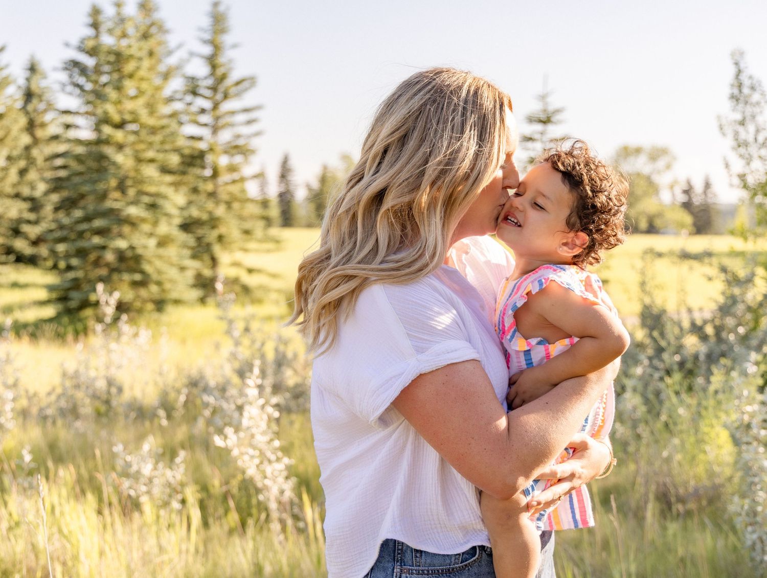 A mother holds and kisses her curly-haired toddler in a sunny green meadow with pine trees in the background.