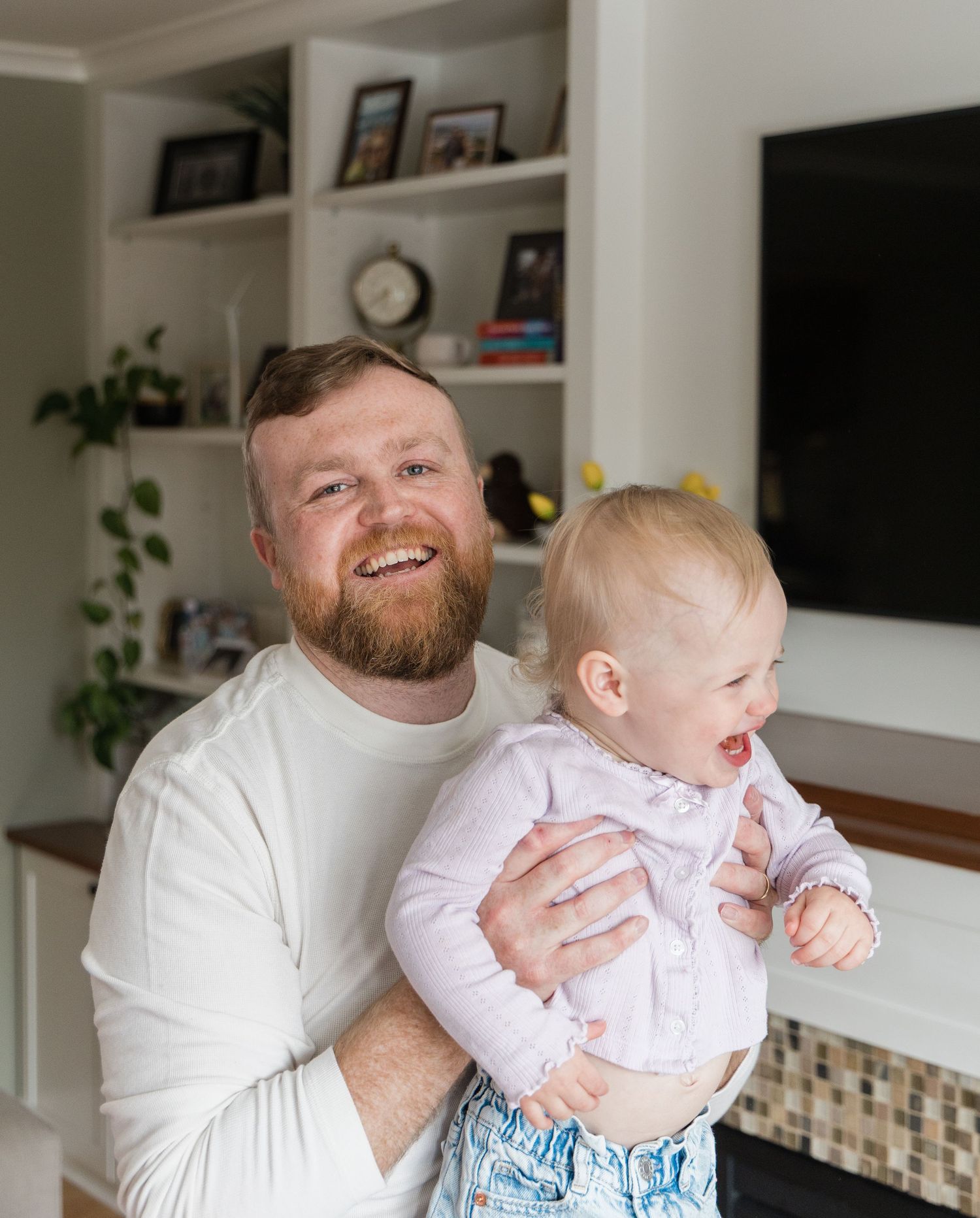 Smiling bearded dad in white shirt holding laughing baby girl in bright living room.
