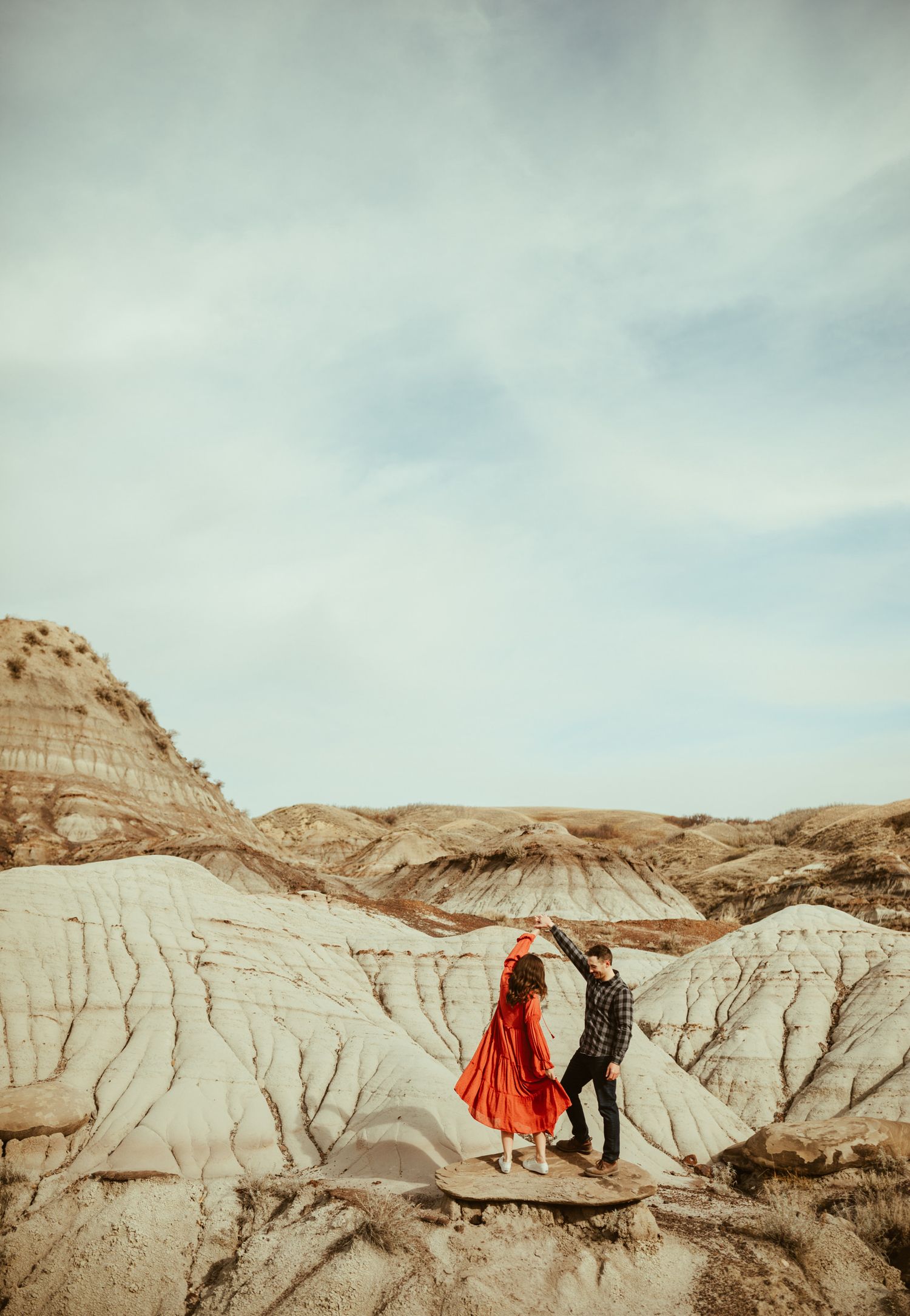 A couple dances on a rocky badlands landscape, the woman wearing a flowing red dress under a cloudy sky.