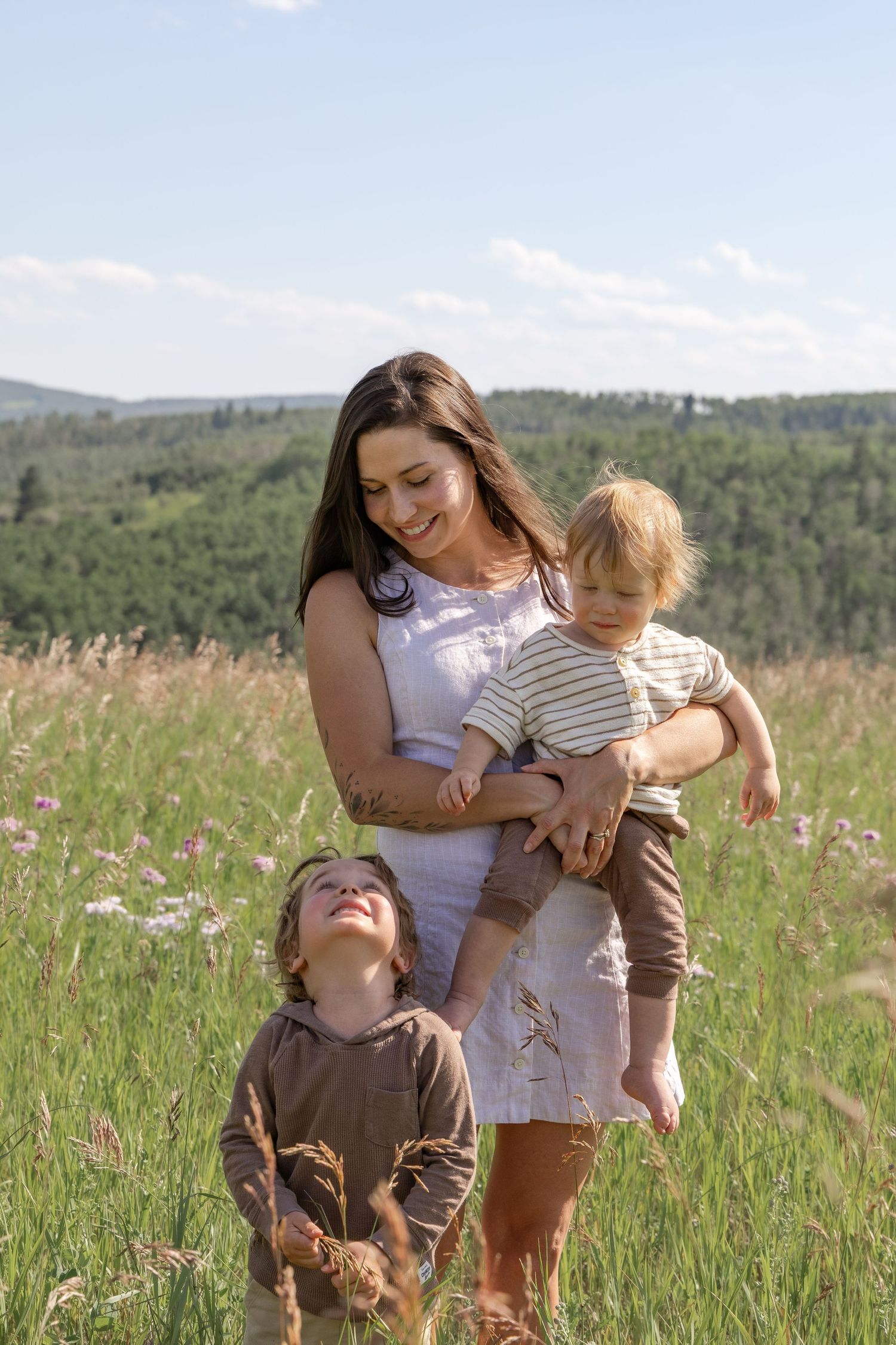 A smiling mother holds a toddler while another young child looks up at her in a sunny green meadow.