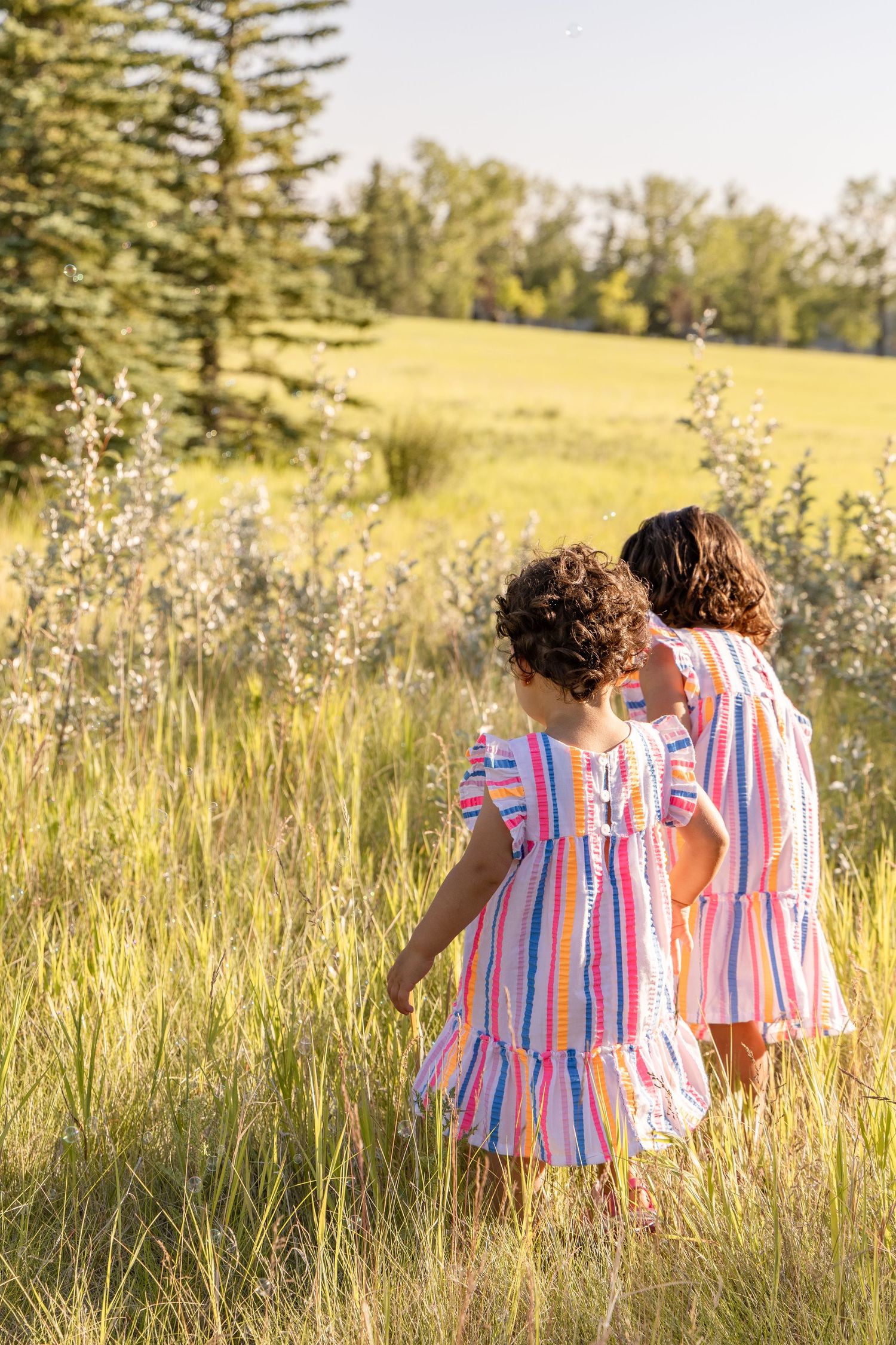 Two young girls in matching colorful striped dresses walk through a sunny green meadow surrounded by tall grass and trees.