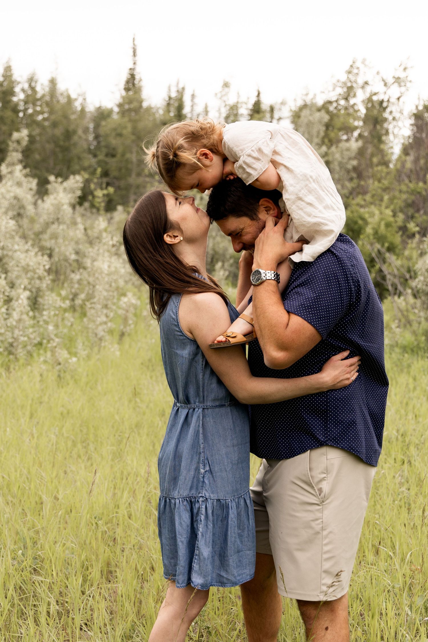 Loving parents lean in for a kiss while dad lifts their young child above them in a green field.