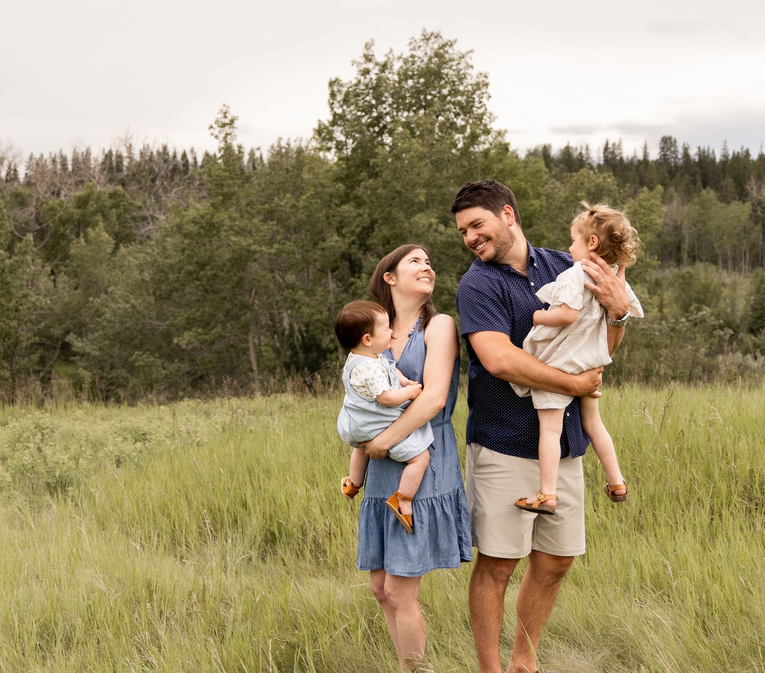 Happy young family of four stands in a lush green meadow, parents each holding a small child.