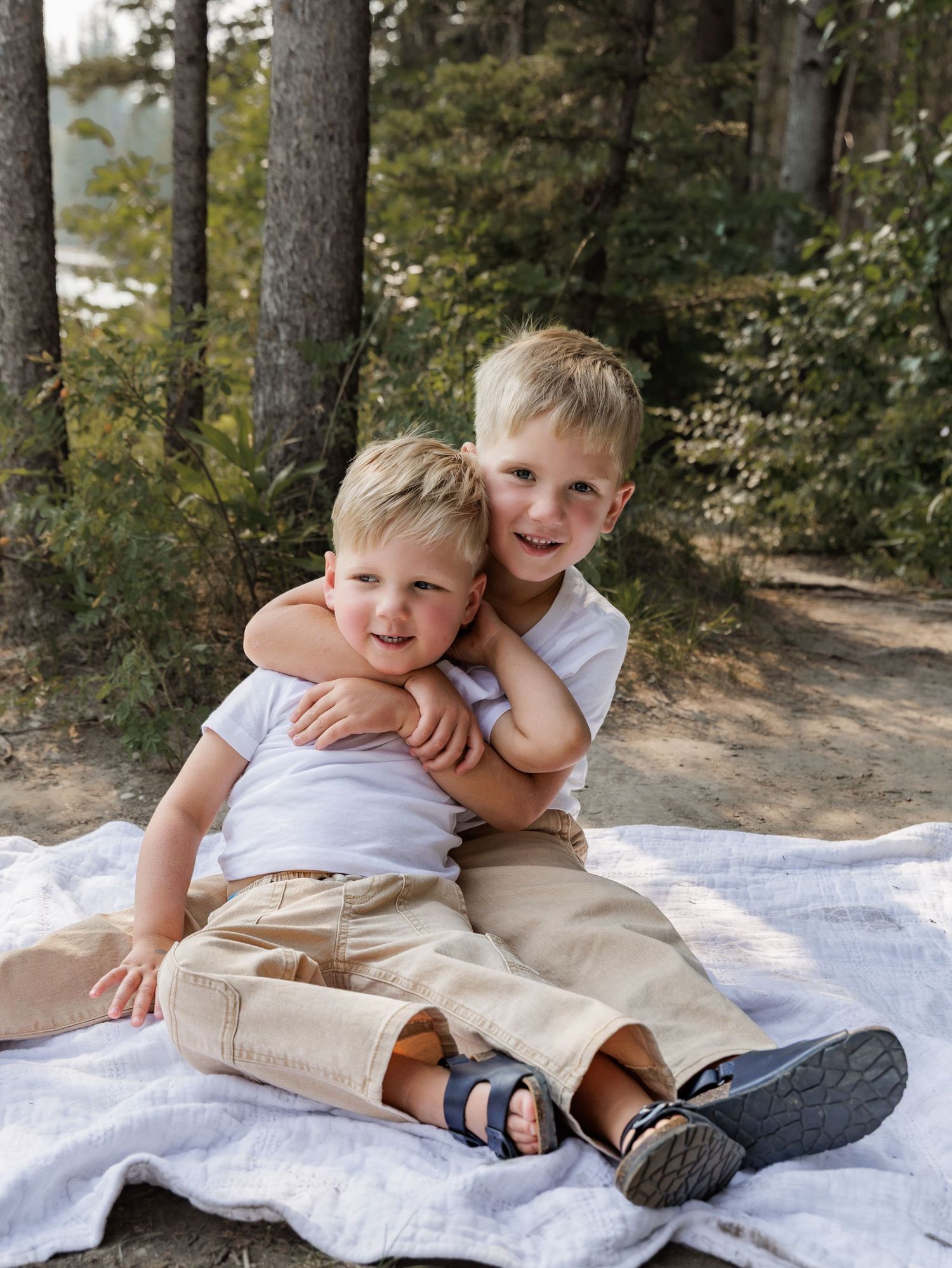 Two young blonde brothers hugging and smiling on a blanket in a forest setting.