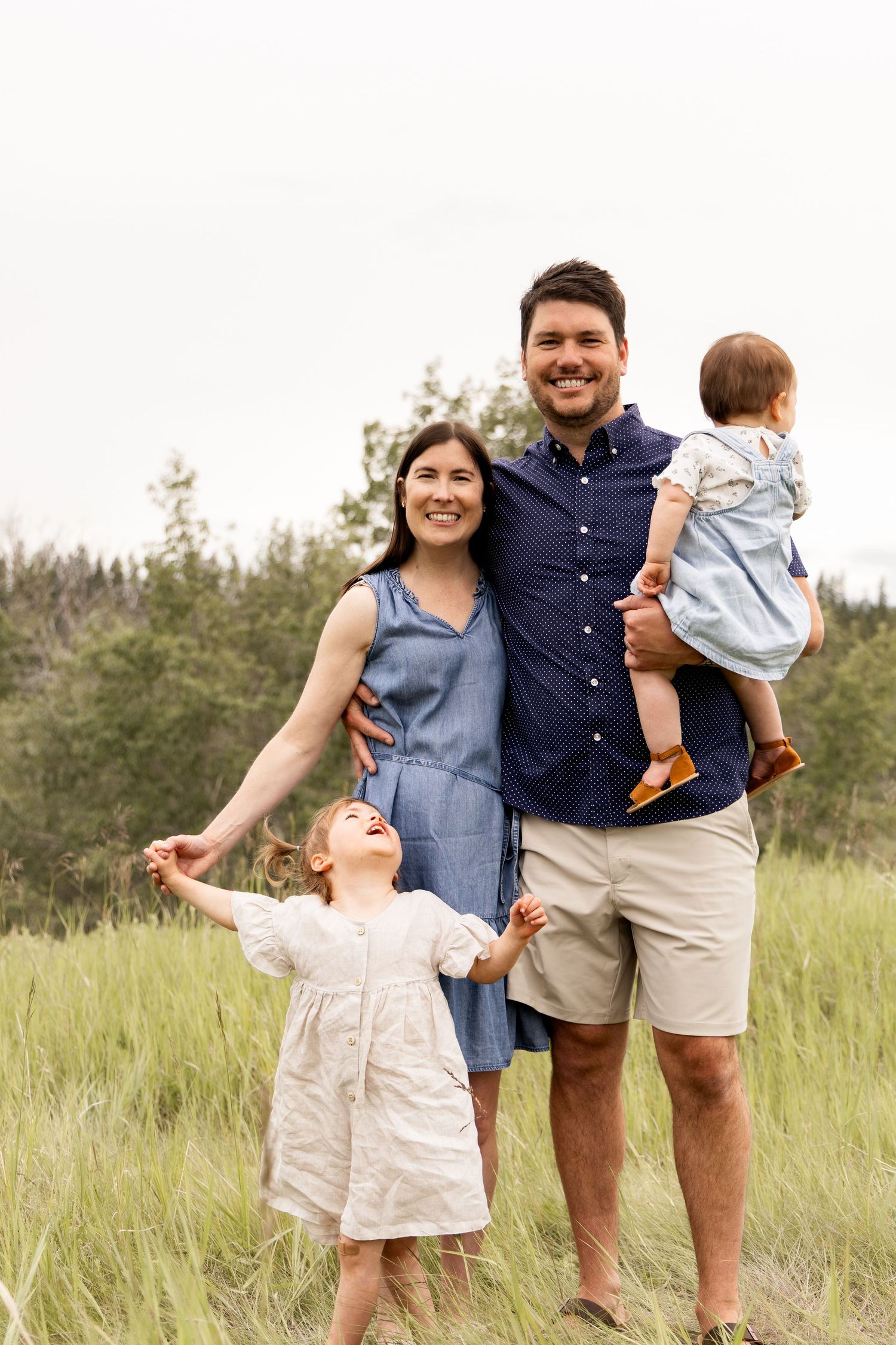 Smiling family of four stands in a green meadow, dad holding baby while toddler daughter reaches up.