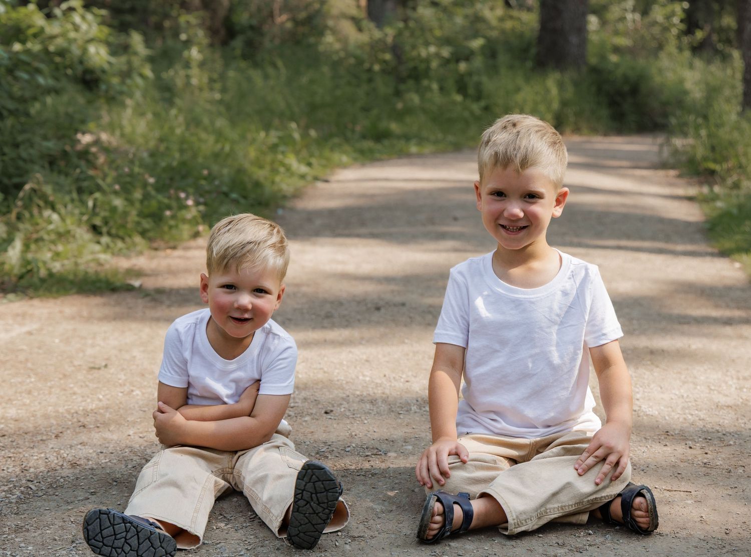 Two smiling blonde boys in white shirts and khaki shorts sitting on a dirt path surrounded by green trees.