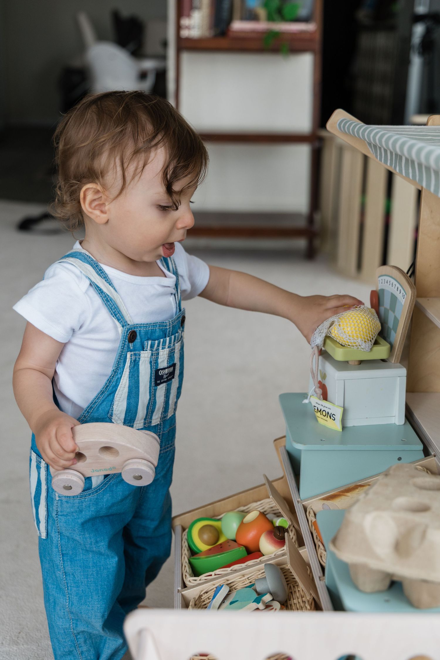 Curious toddler in denim overalls playing with wooden toys at a play kitchen.