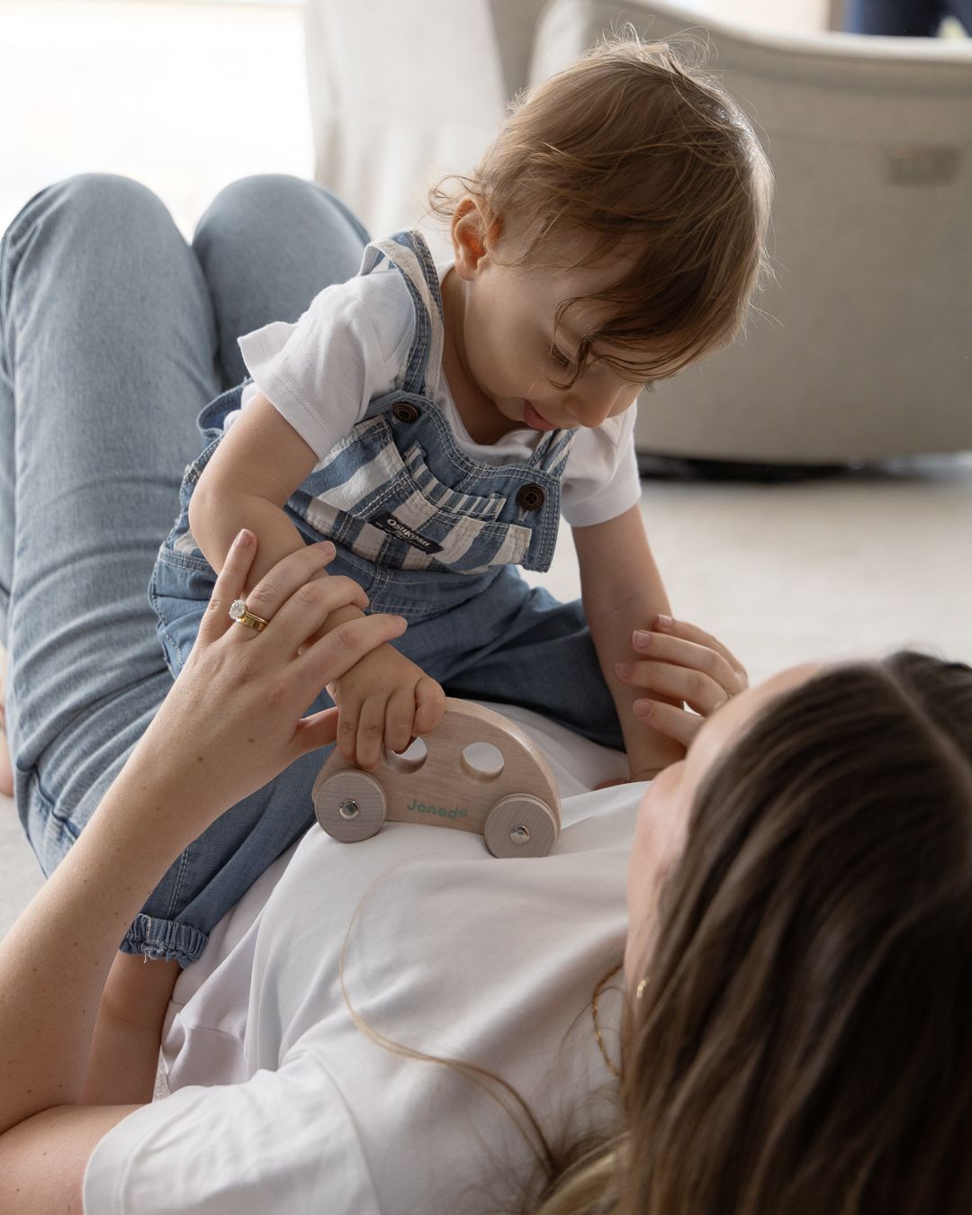 A mother lying on the floor playing with her toddler who holds a wooden toy car indoors.