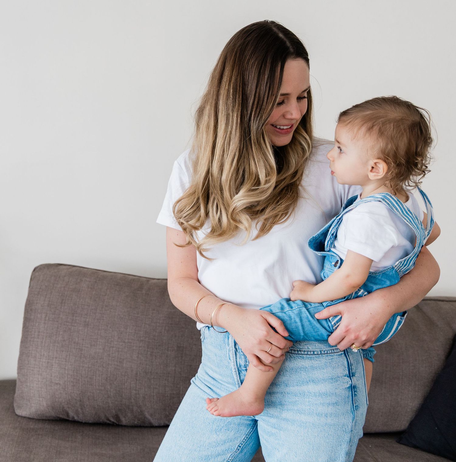 Smiling mother in white tee holding toddler in denim overalls while sitting on a gray sofa.