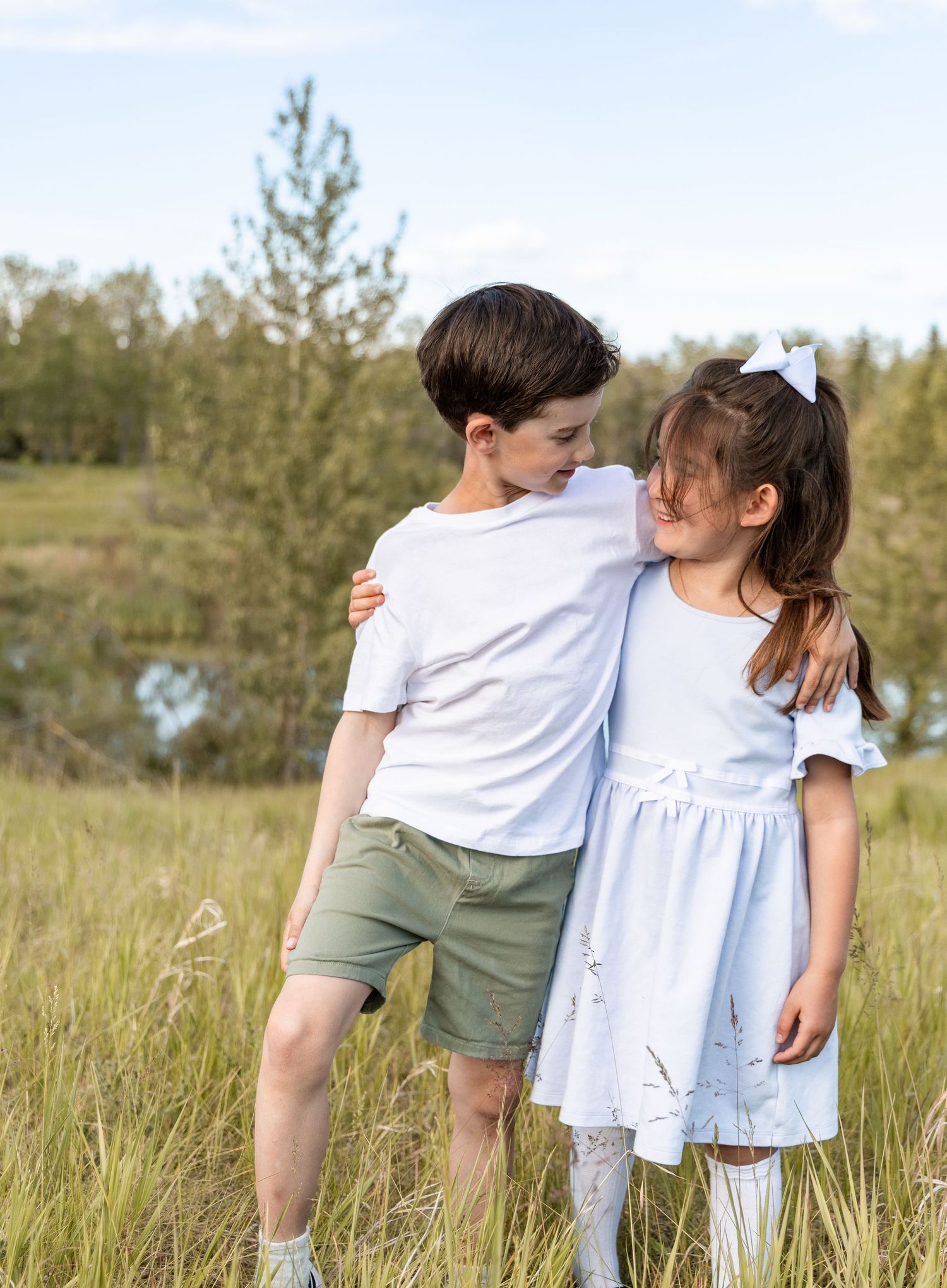 Two children hugging in a grassy field, boy in white tee and green shorts, girl in light blue dress with white bow.