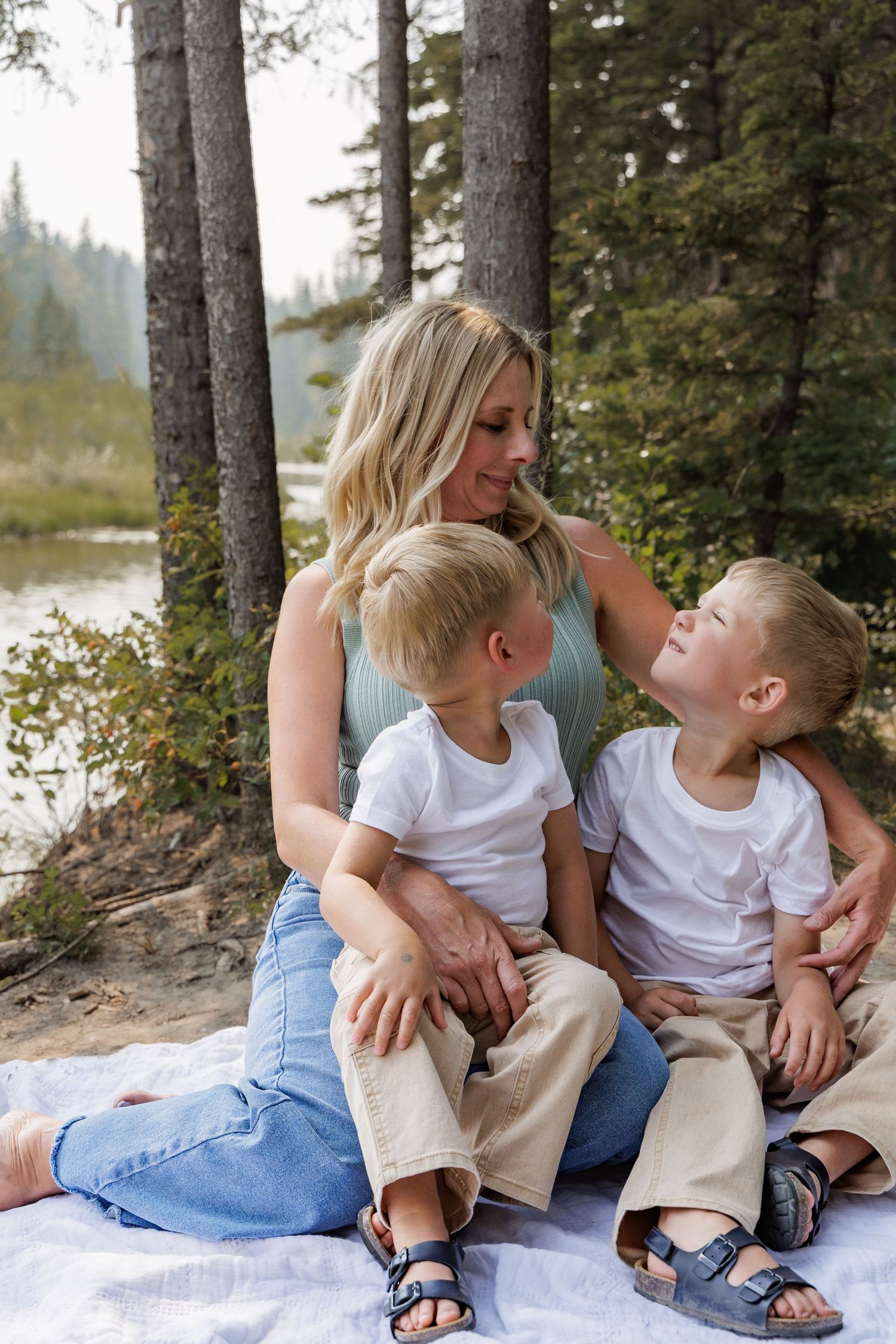 A blonde mother sits on a blanket with her two young sons near a forest lake, smiling warmly at each other.