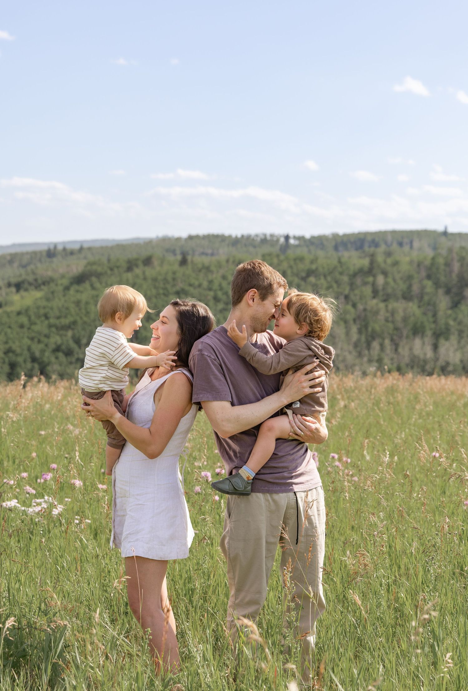 A happy family of four poses in a sunny green meadow, parents holding two young children with forest hills behind.
