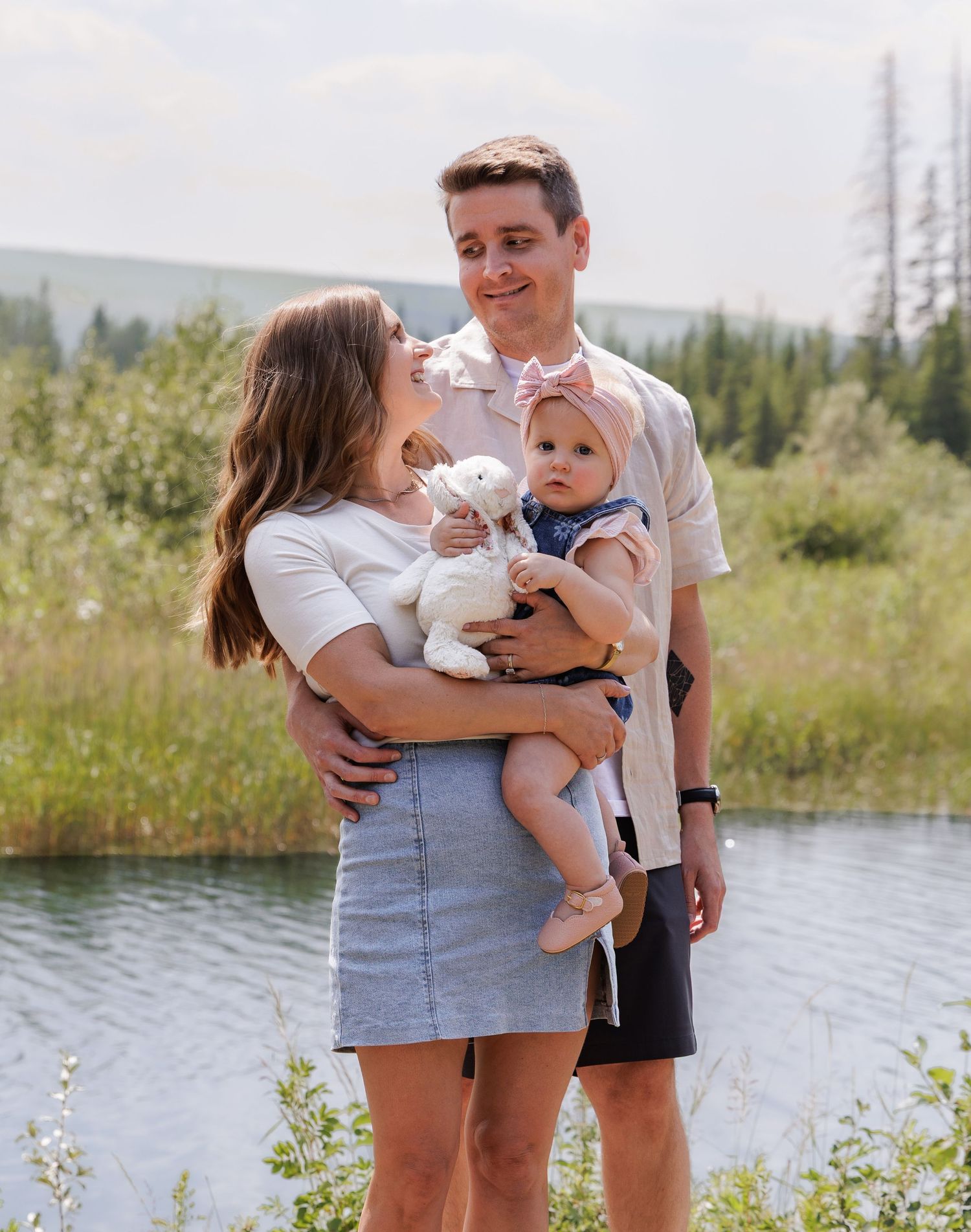 Young couple holding baby with stuffed animal poses outdoors near lake surrounded by lush green forest.