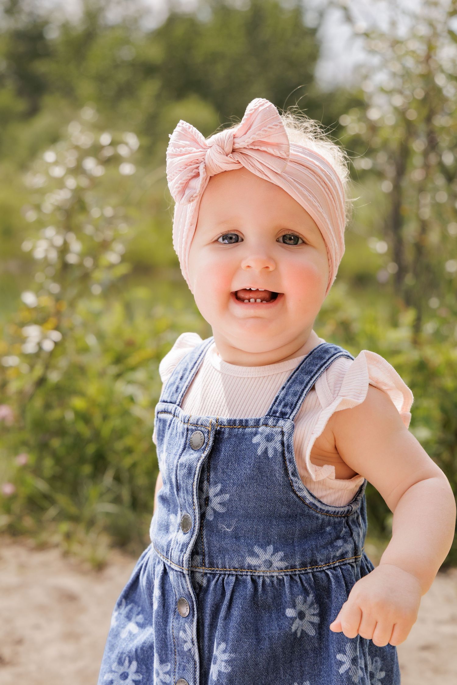 Smiling toddler girl in denim floral overalls and pink bow headband standing outdoors in a sunny garden.
