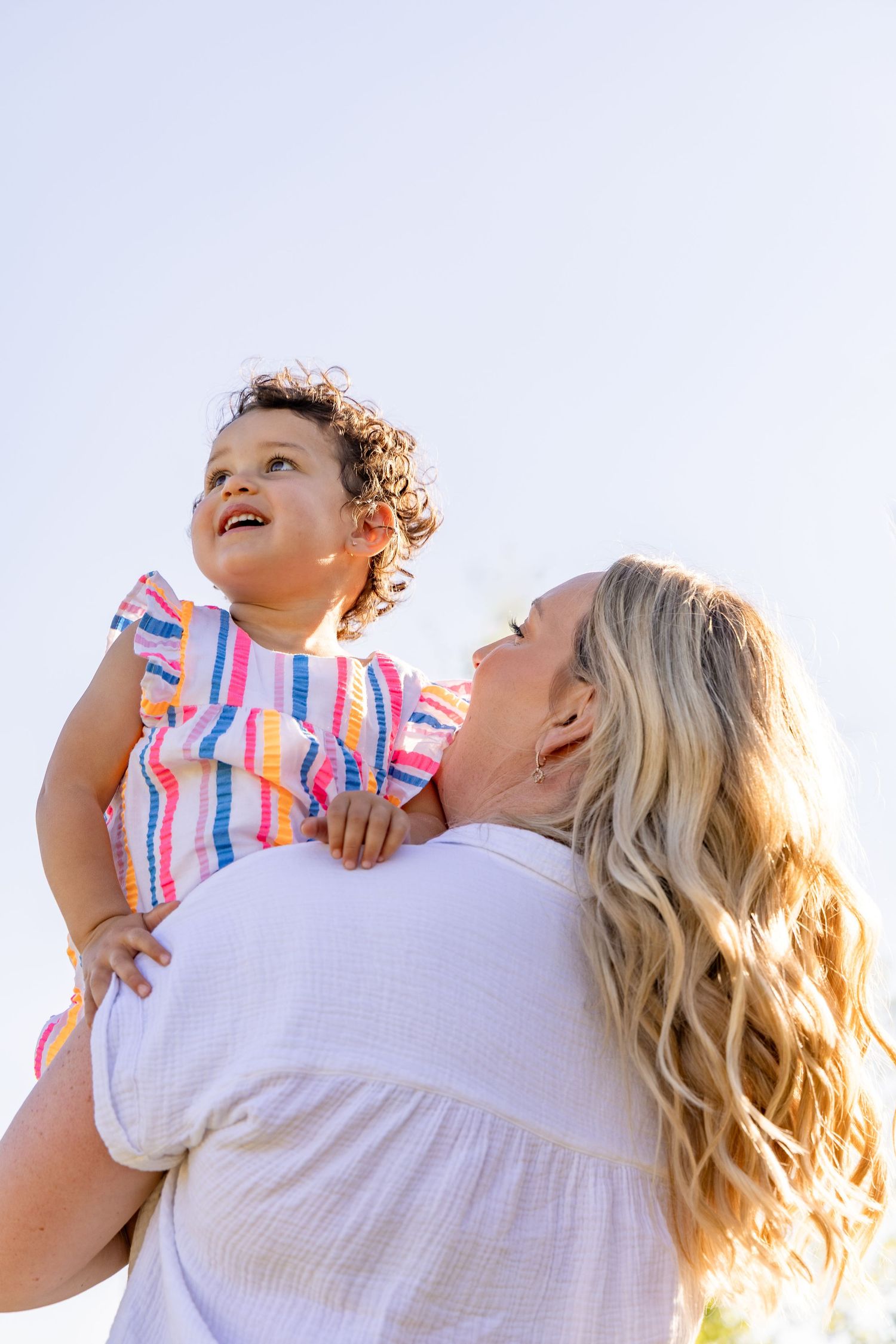 A blonde woman lovingly holds a curly-haired toddler in a colorful striped dress against a bright sky.