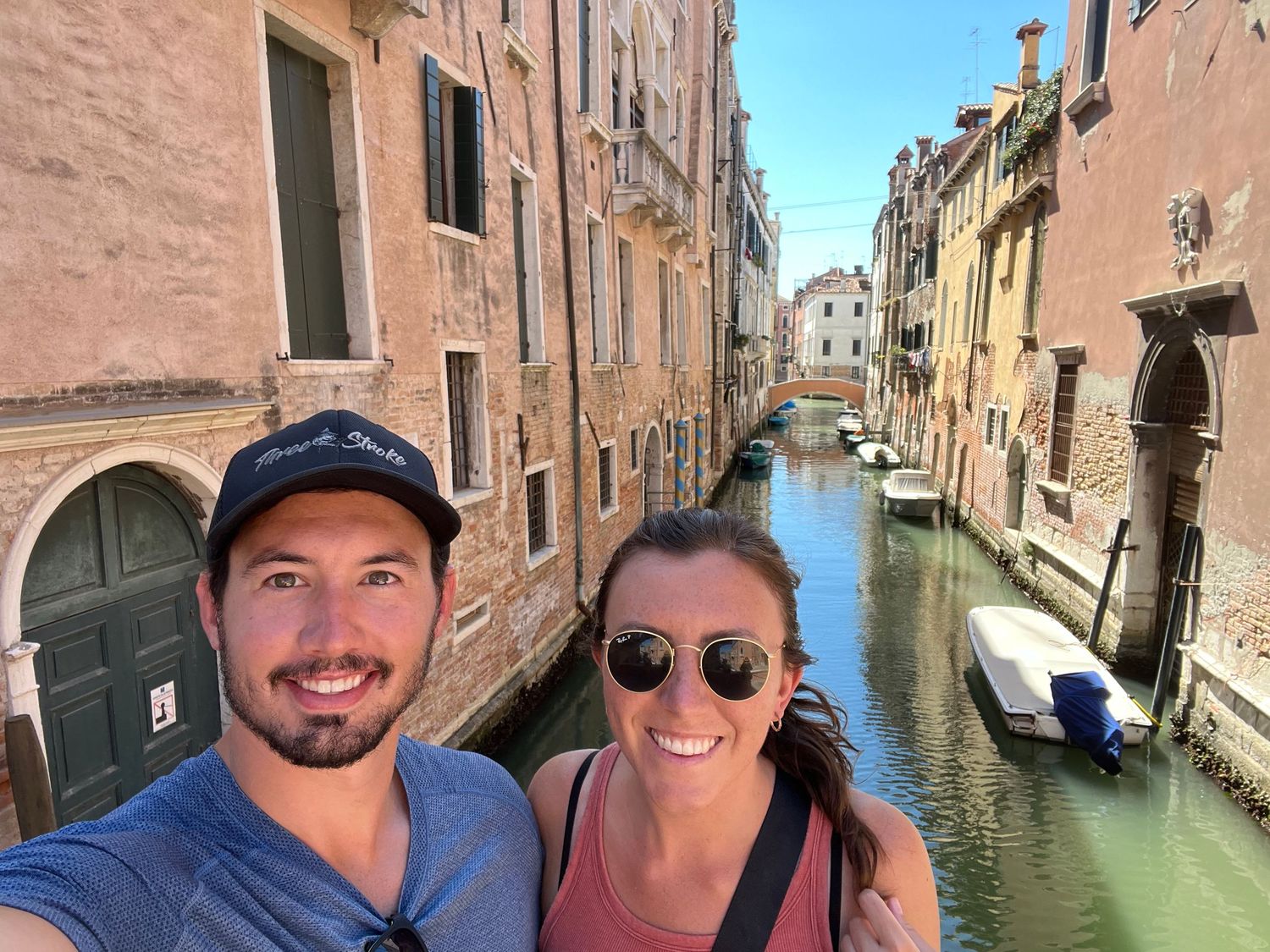 A smiling couple takes a selfie on a bridge over a scenic Venice canal lined with historic buildings.