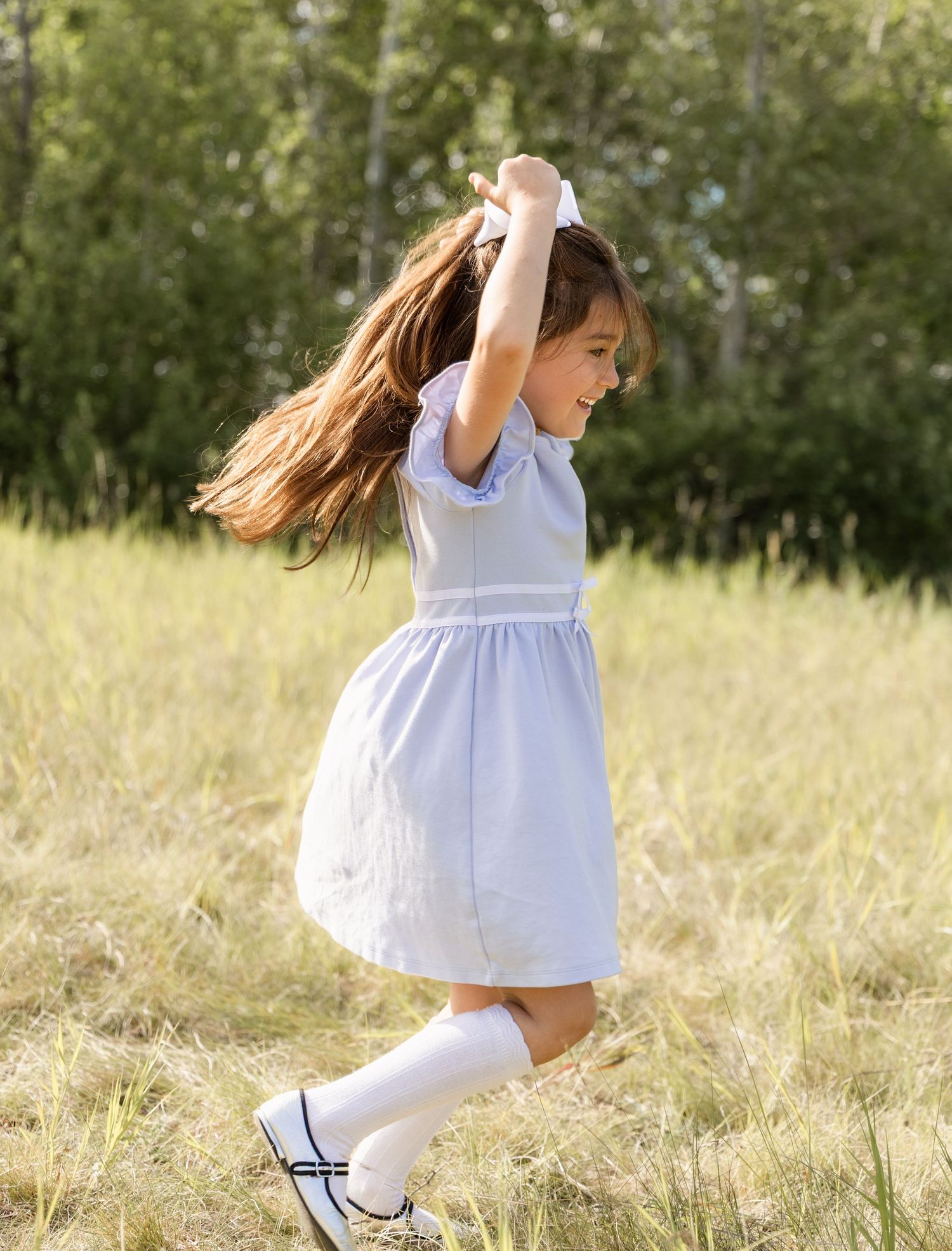 A young girl in a light blue dress twirls joyfully in a sunny golden meadow surrounded by trees.