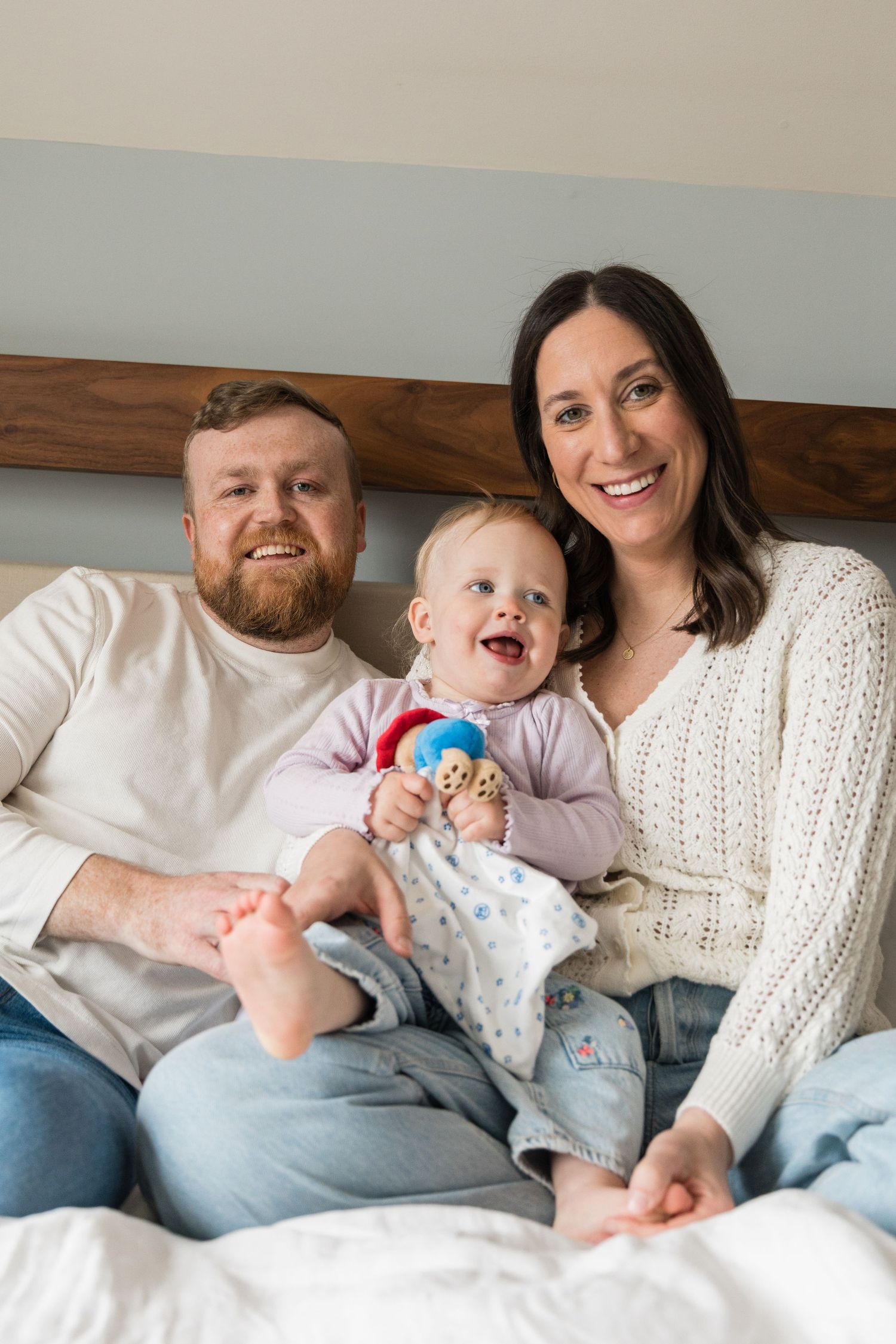 Happy family portrait with bearded dad, brunette mom in white sweater, and laughing baby holding colorful toy on bed.