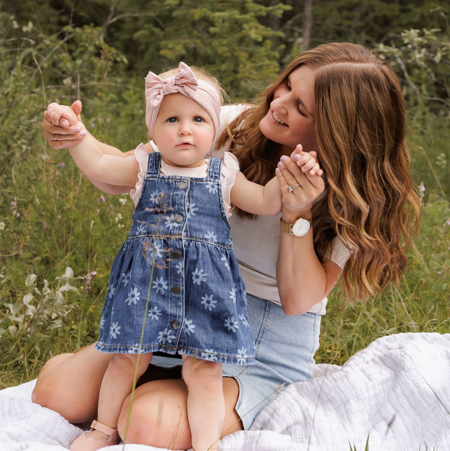 A smiling mother sits on a blanket outdoors helping her baby girl in a denim dress and pink bow stand up.