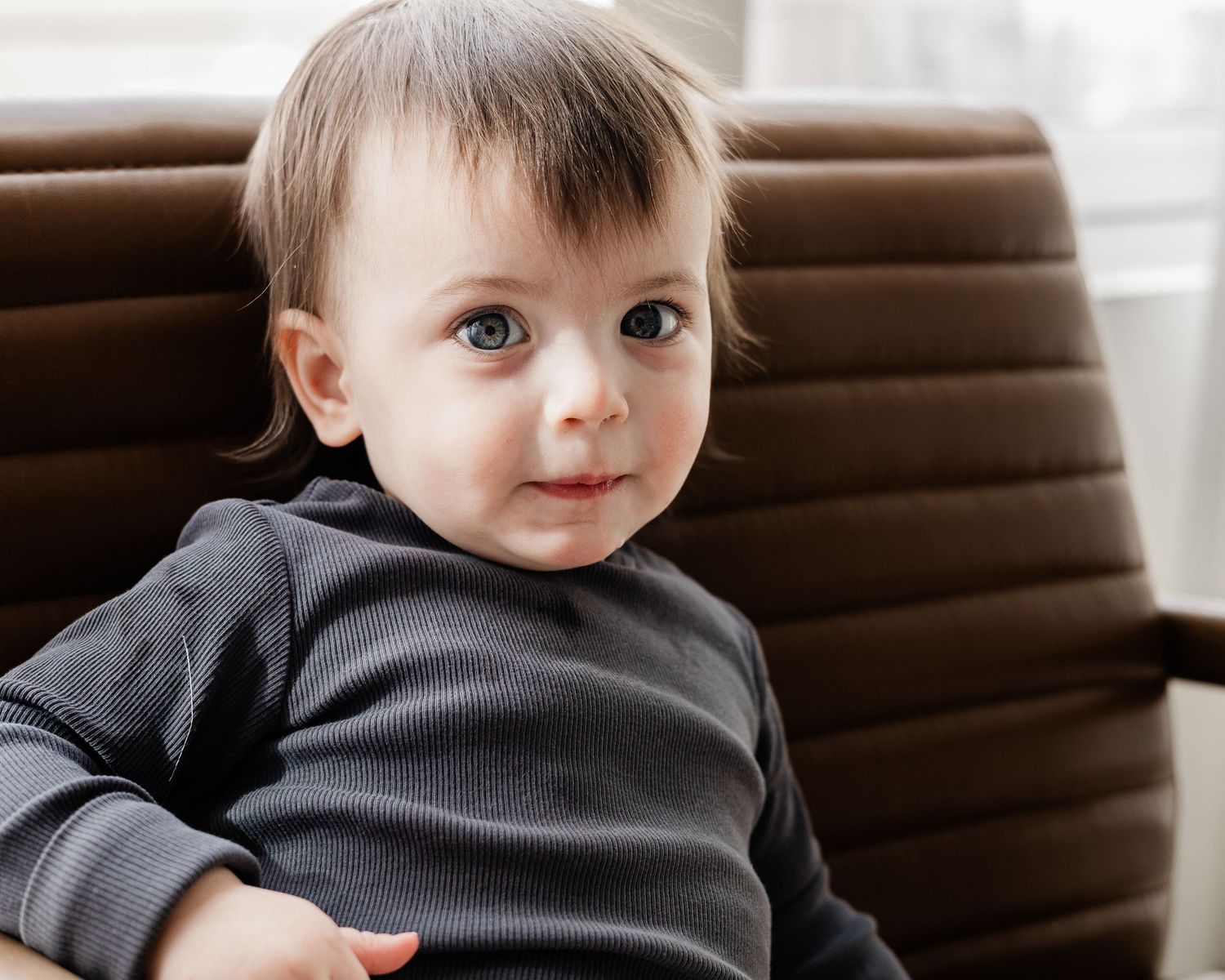 Adorable baby boy with dark hair sitting in a brown leather chair looking at camera.