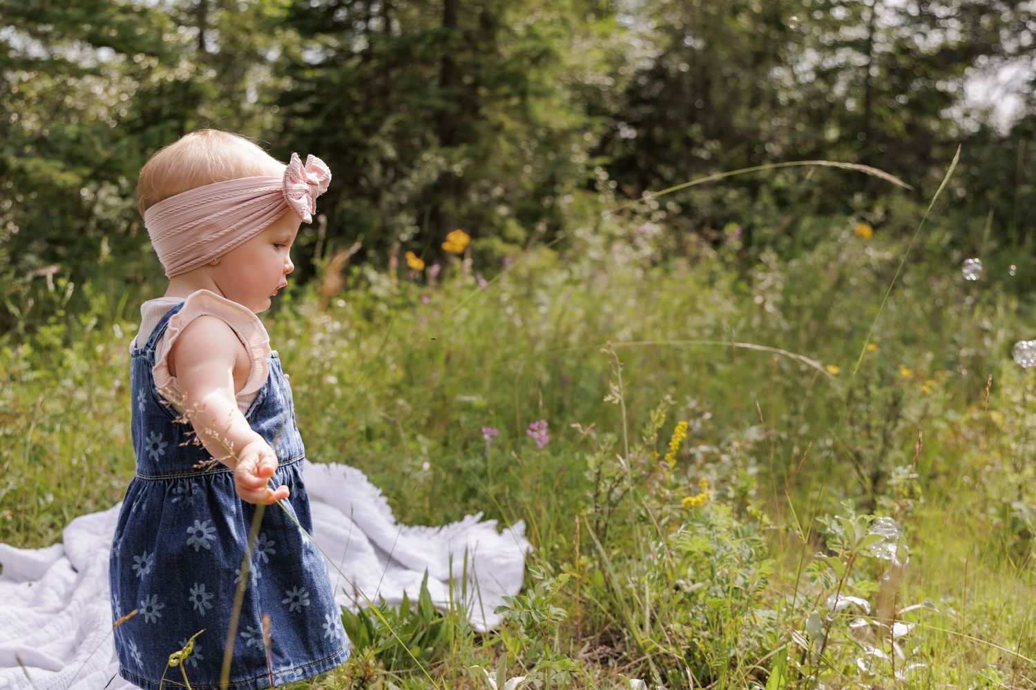 A toddler girl in a blue floral dress and pink headband plays in a lush green meadow on a white blanket.