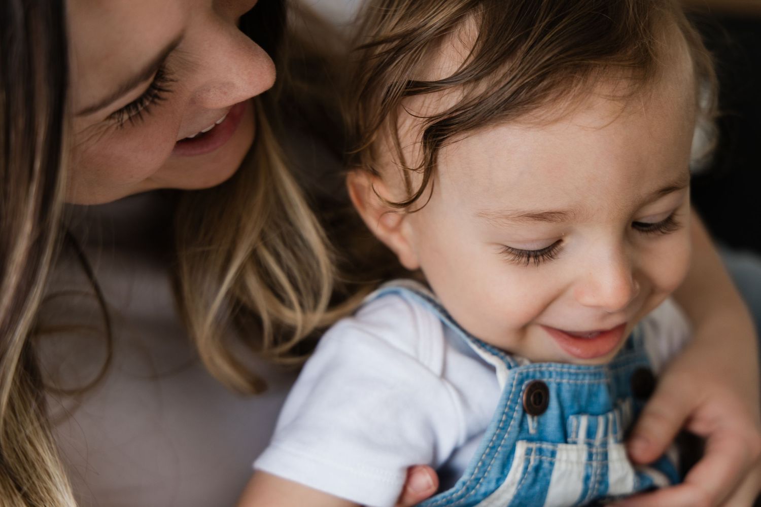 Close-up of a smiling toddler in denim overalls being held by their mother.