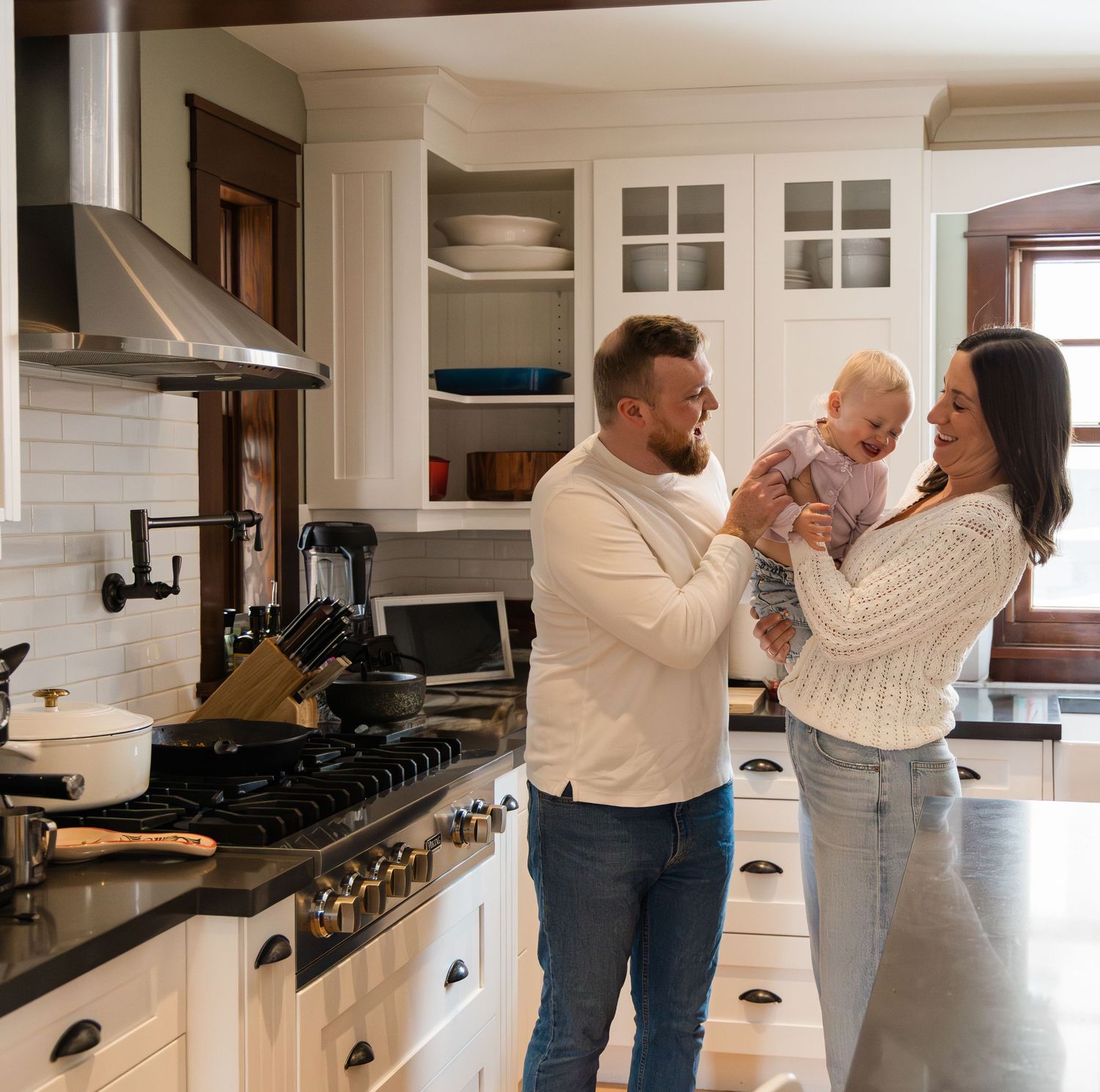 Smiling couple in white sweaters and jeans holding baby girl together in bright white kitchen.