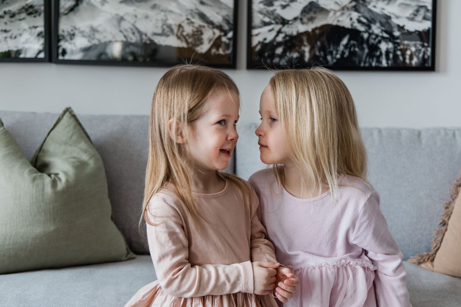 Two adorable blonde sisters in pink dresses sitting on a gray sofa, giggling and hugging each other.